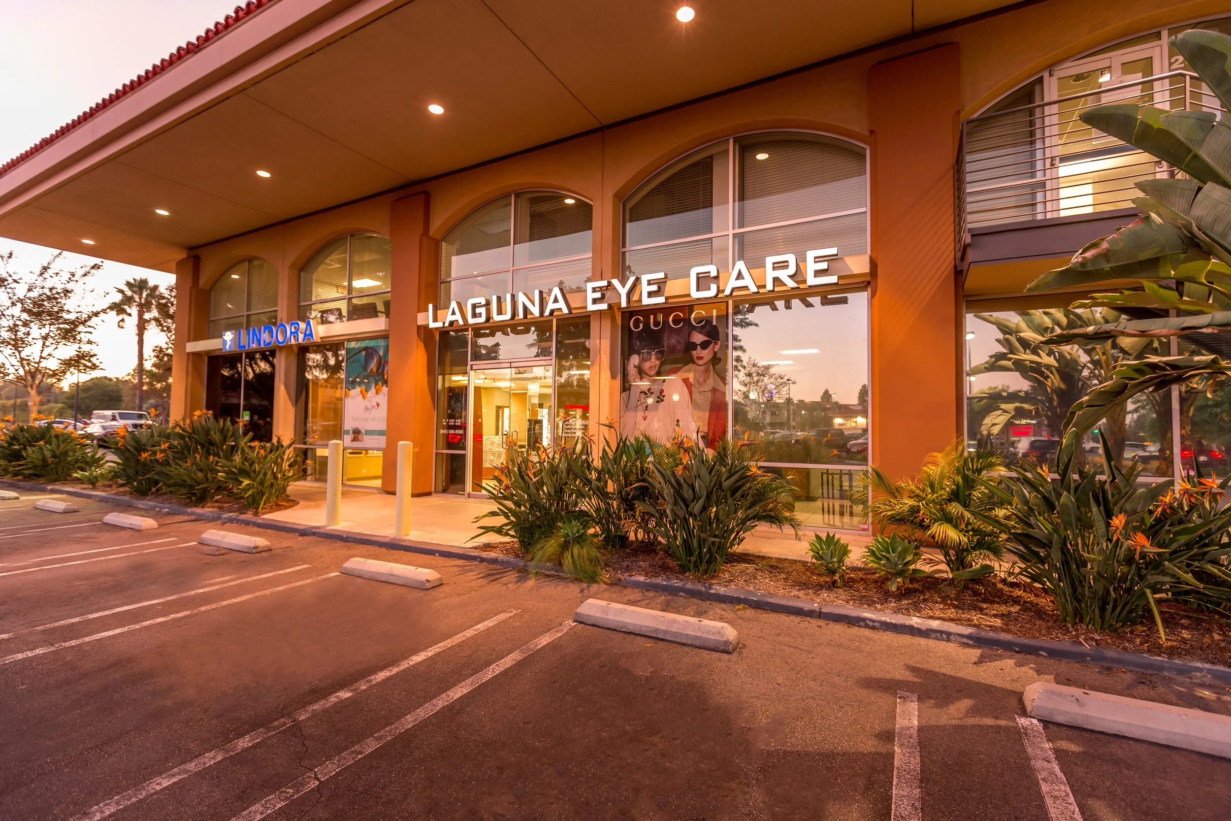 View of a shopping center with stores named Lindora and Laguna Eye Care, featuring large windows, a parking lot with empty parking spaces, and decorative plants outside, during sunset.