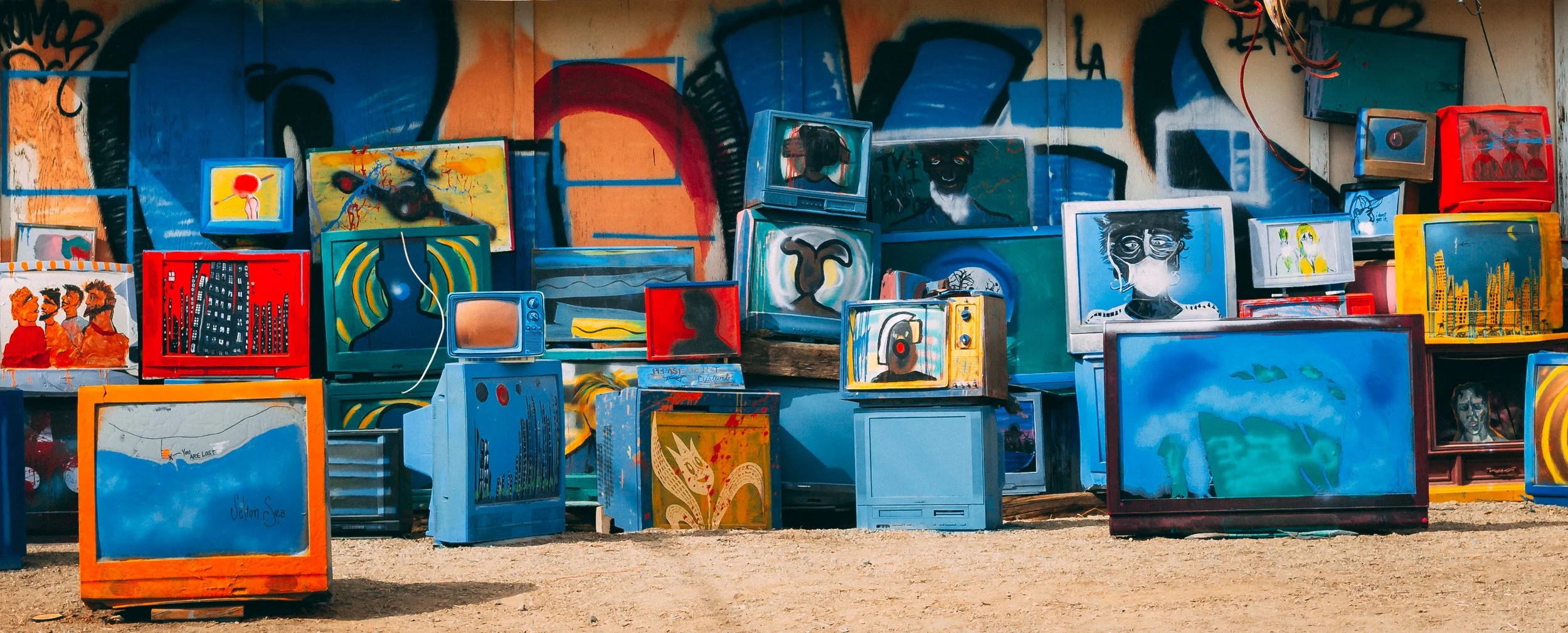 A collection of vintage and modern televisions with colorful and abstract artwork displayed on their screens, arranged outdoors against a graffiti-covered wall.