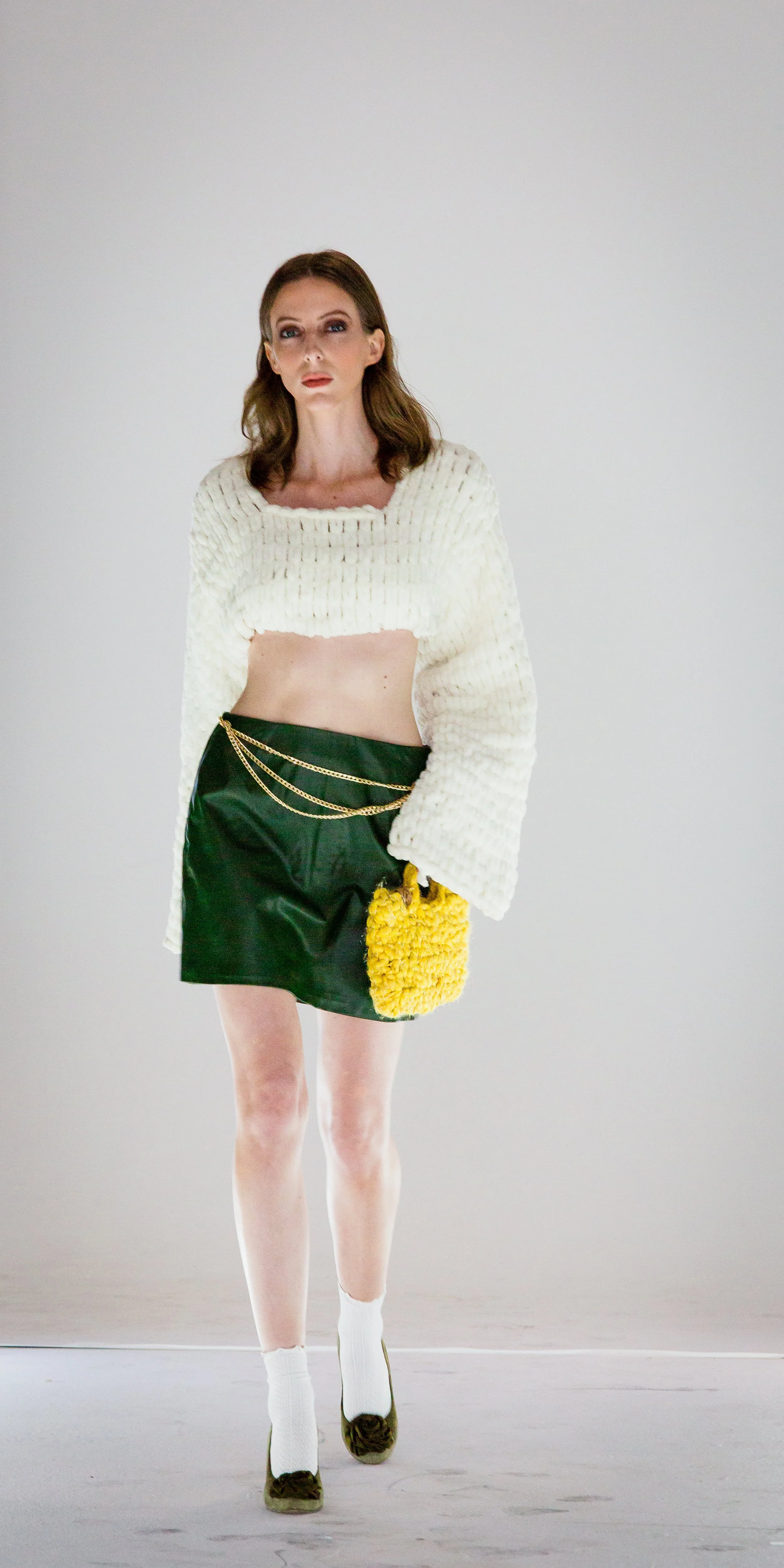 A woman with brown hair and light skin wearing a white knitted cropped sweater, dark green skirt, white socks, and olive green shoes, holding a yellow fuzzy handbag, standing against a plain background.