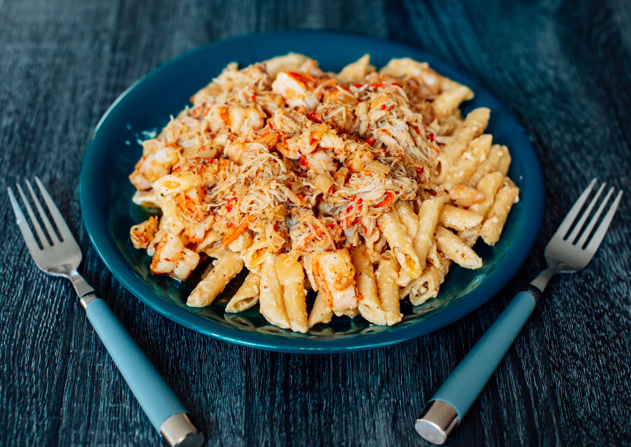 A plate of pasta with shredded chicken and red pepper flakes on top, served on a dark wooden table with two forks on either side.