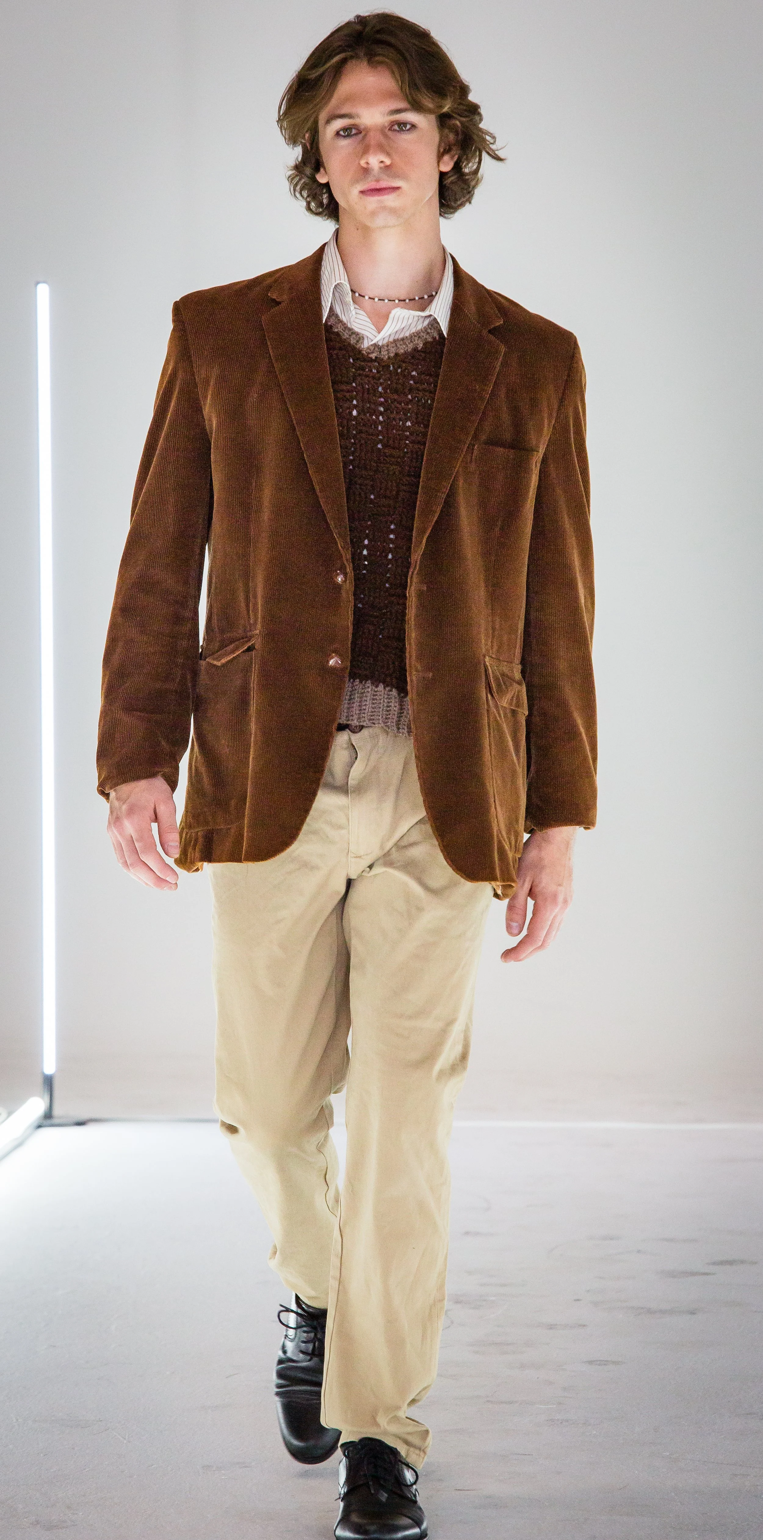 A young man with wavy brown hair wearing a brown velvet blazer over a striped shirt, layered with a dark brown sweater, beige pants, and black shoes, walking in a plain, well-lit environment.