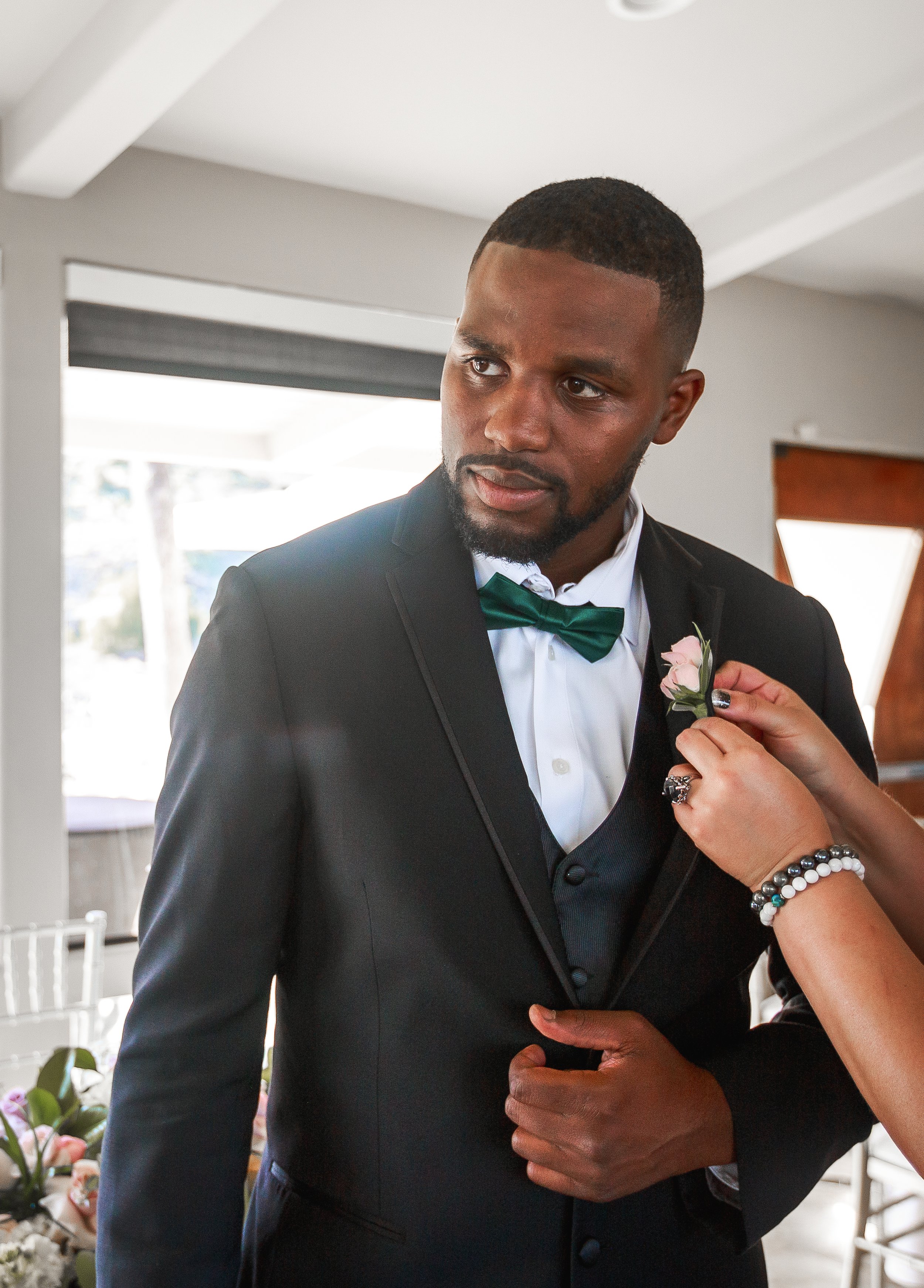 A man in a black tuxedo with a green bow tie is getting a pink boutonniere pinned on his lapel by a woman, during what appears to be a wedding or formal event.