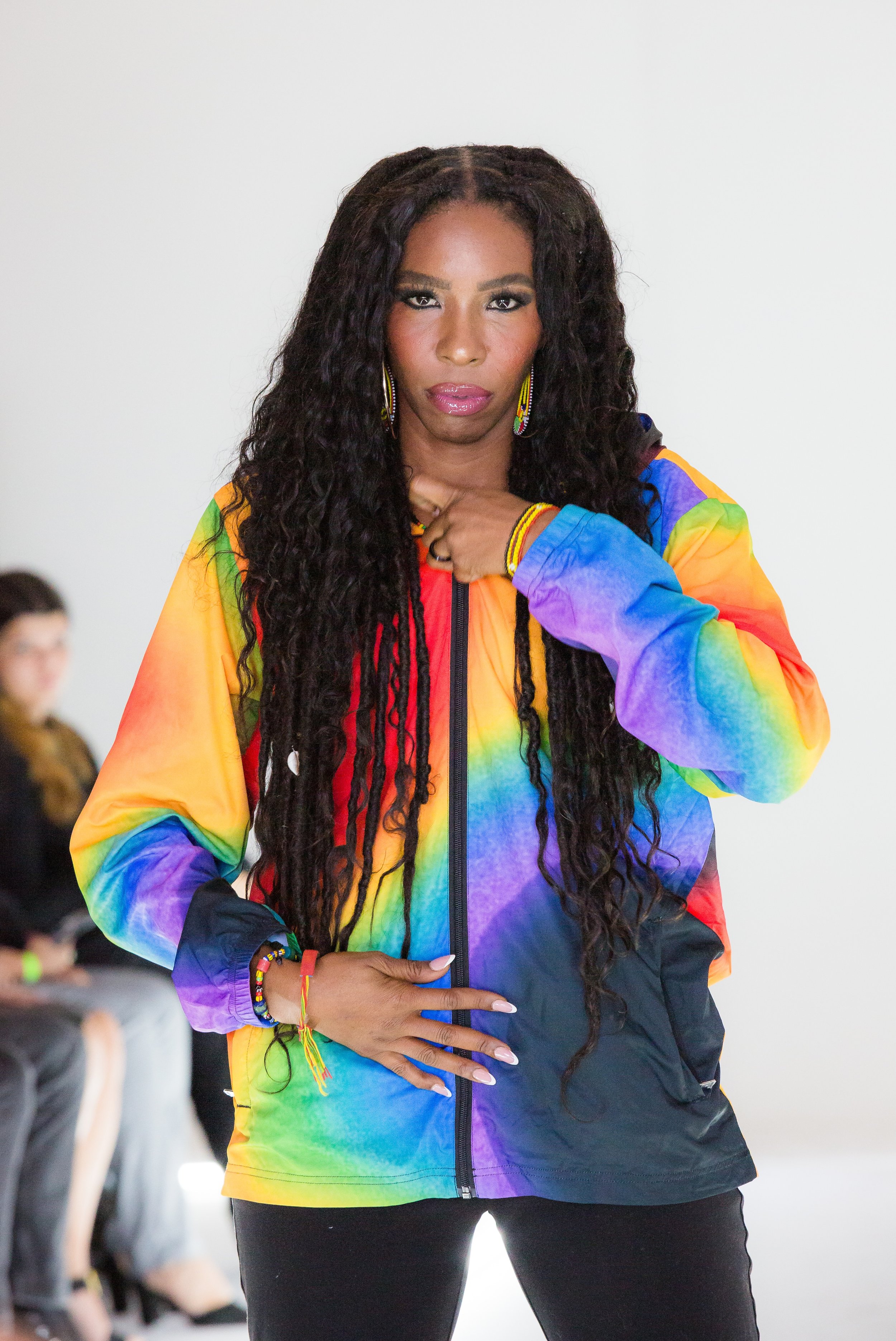Woman with long curly hair wearing a vibrant rainbow-colored jacket, standing indoors with a neutral background.
