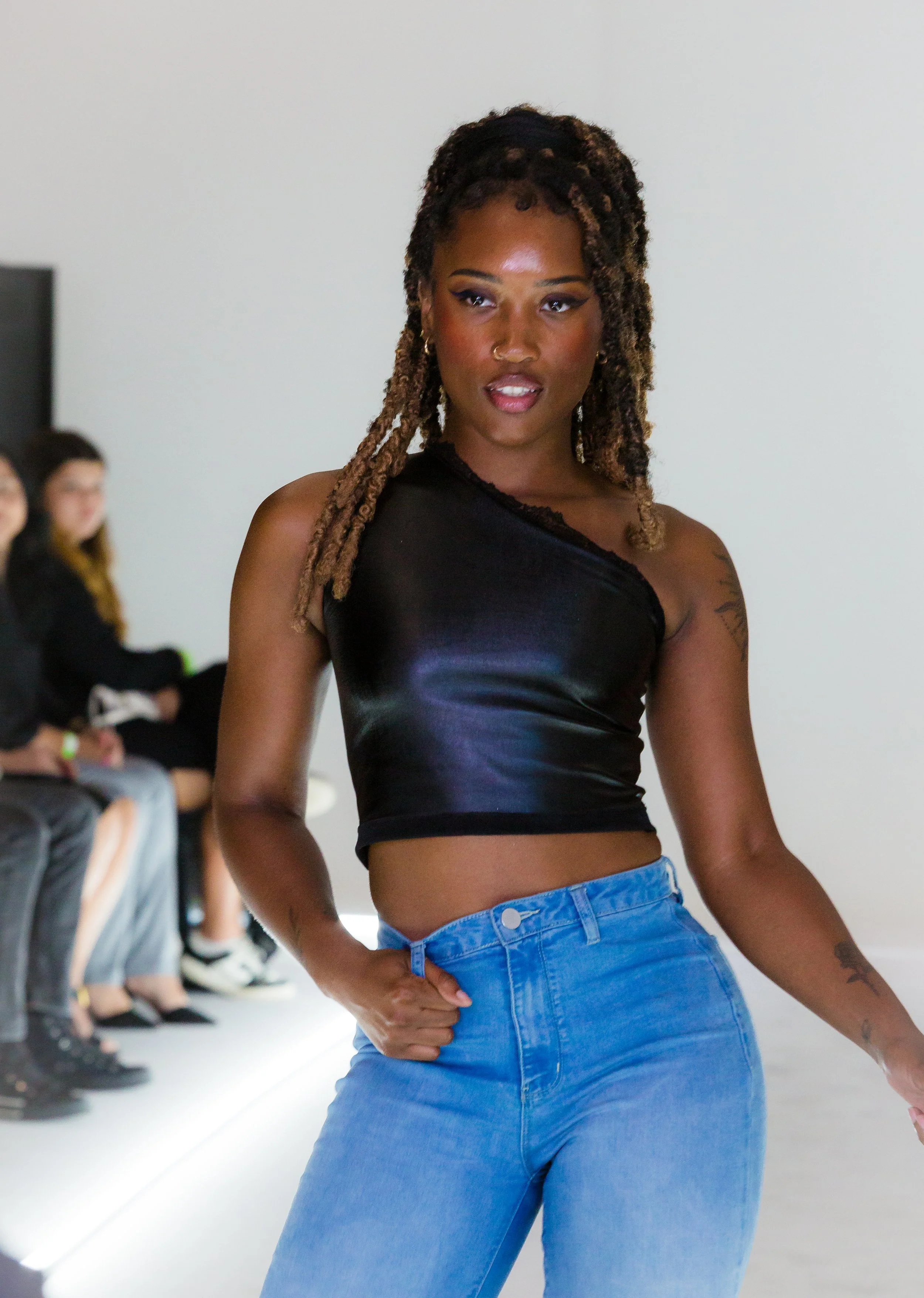 A woman with styled dreadlocks walking on a runway, wearing a black shiny sleeveless crop top and blue high-waisted jeans, with a small nose ring and tattoos on her arms.