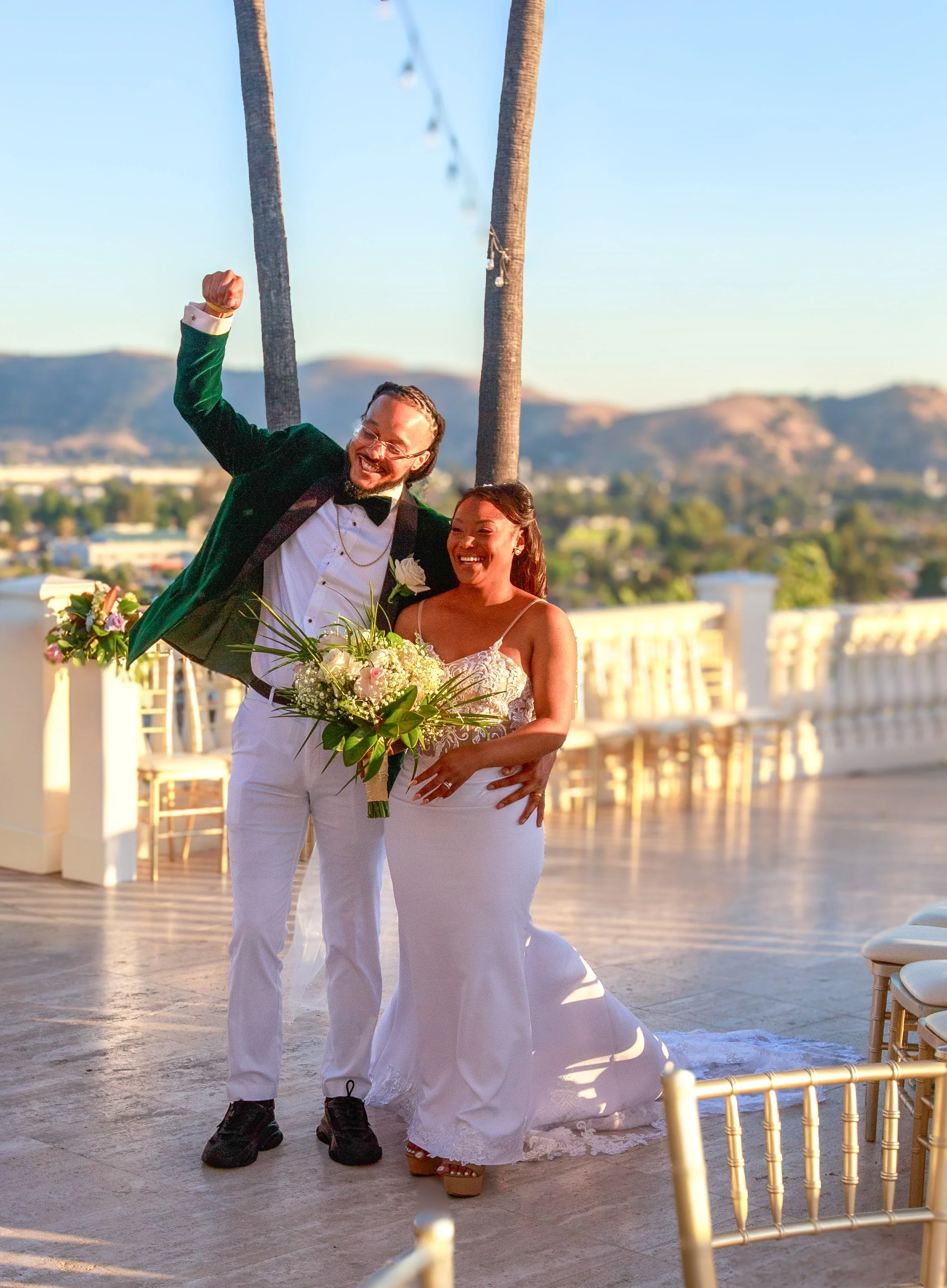 A newlywed couple enjoying their wedding celebration outdoors during sunset, with a scenic mountain view in the background. The groom is in a tuxedo and the bride in a white wedding gown holding a bouquet of flowers.