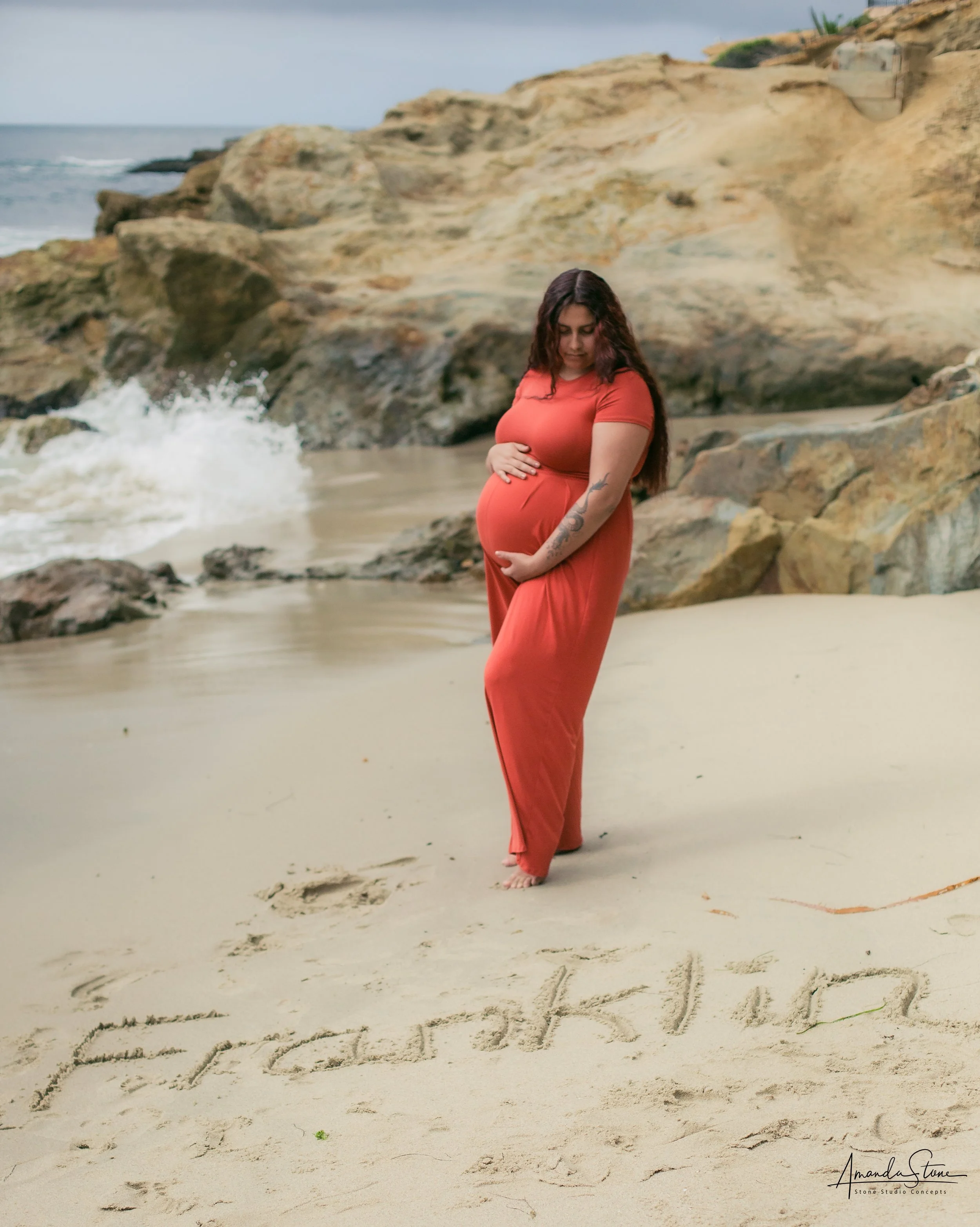 Pregnant woman in orange dress standing on beach, holding her belly and looking down, with the name 'Erick' and other writing in the sand.