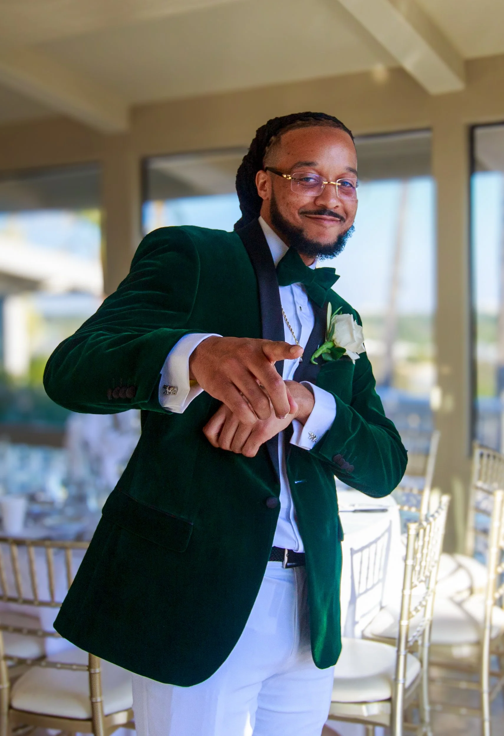 A man dressed in a green velvet blazer, white shirt, and black bow tie, with glasses and a boutonniere, smiling and pointing at the camera in a bright indoor setting with large windows and chairs in the background.