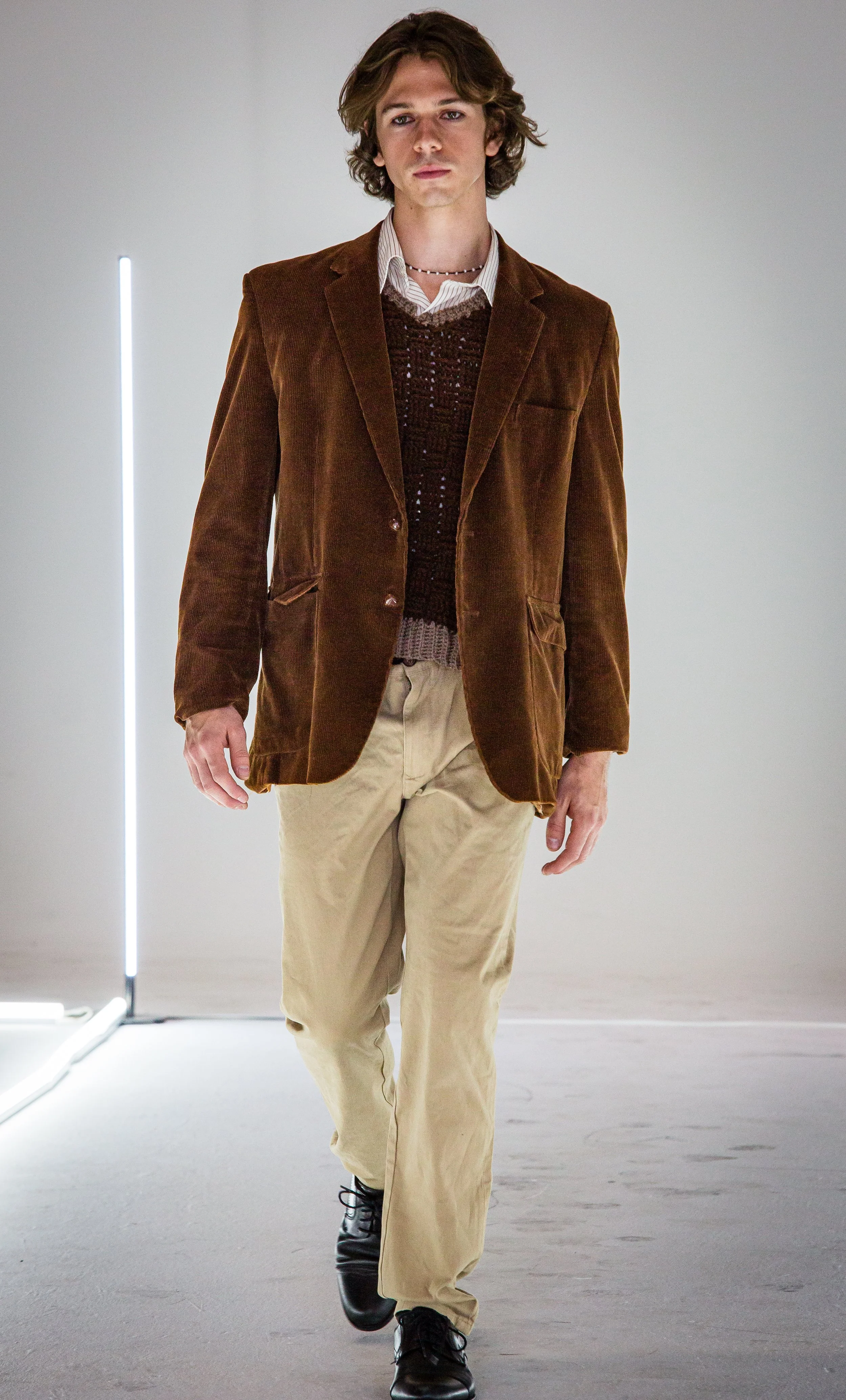 A young man with wavy brown hair walking on a runway, wearing a brown blazer, beige pants, and black shoes, with a plain light background and bright lighting.