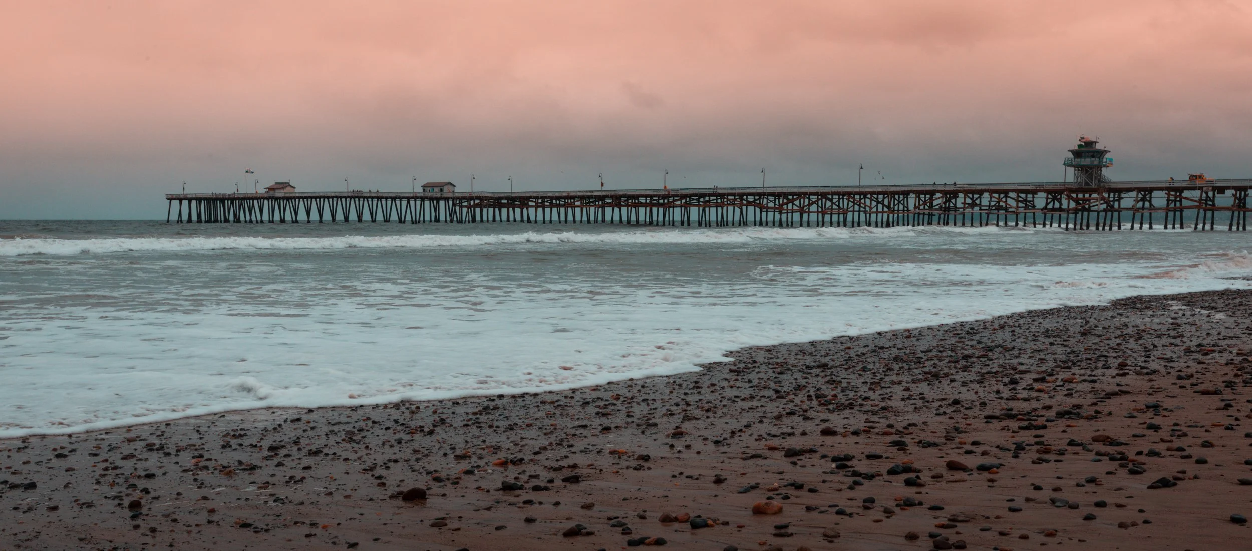A wooden pier extending into the ocean with a lighthouse structure on the right end, under a pinkish-gray sky, near a pebble-covered beach.
