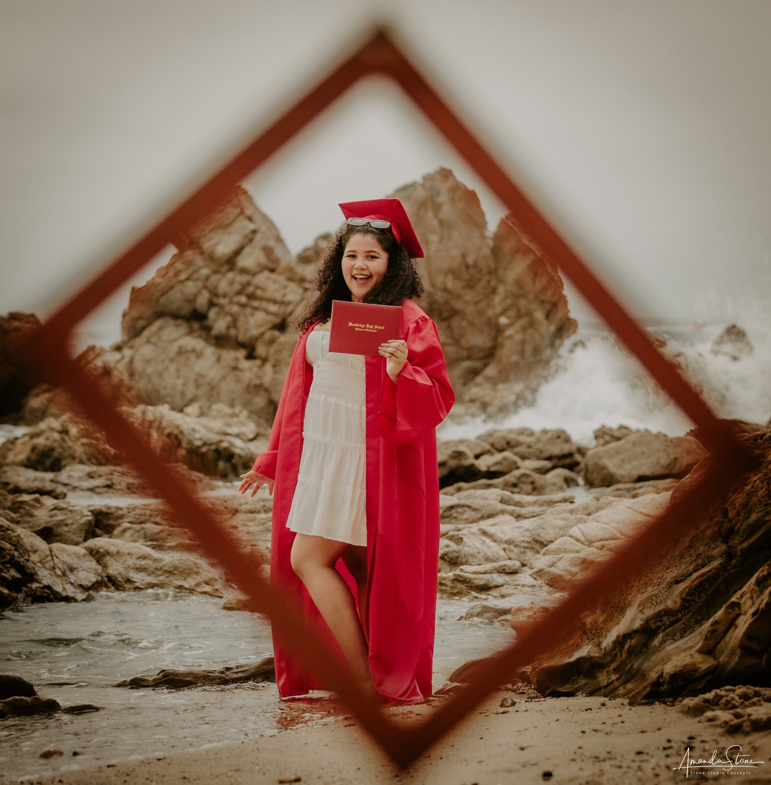A young woman in a graduation cap and gown holding a diploma, smiling and posing on a rocky beach with large rocks and ocean waves in the background, framed by a diamond-shaped border.