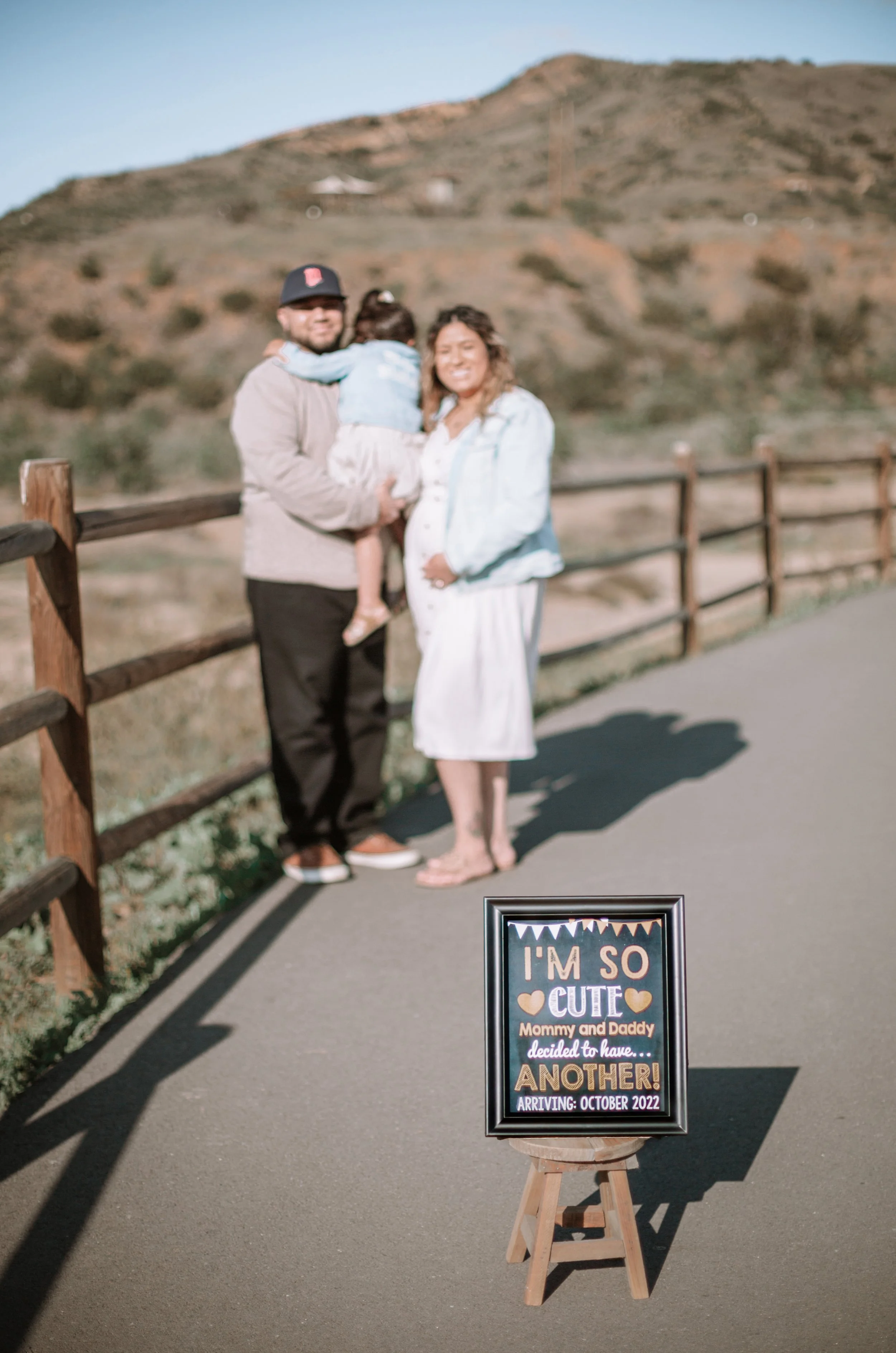 A family of three standing outdoors near a wooden fence, with a sign on a small easel in the foreground that reads 'I'm so cute Mommy and Daddy decided to have... another! Arriving: October 2022', indicating a pregnancy announcement.