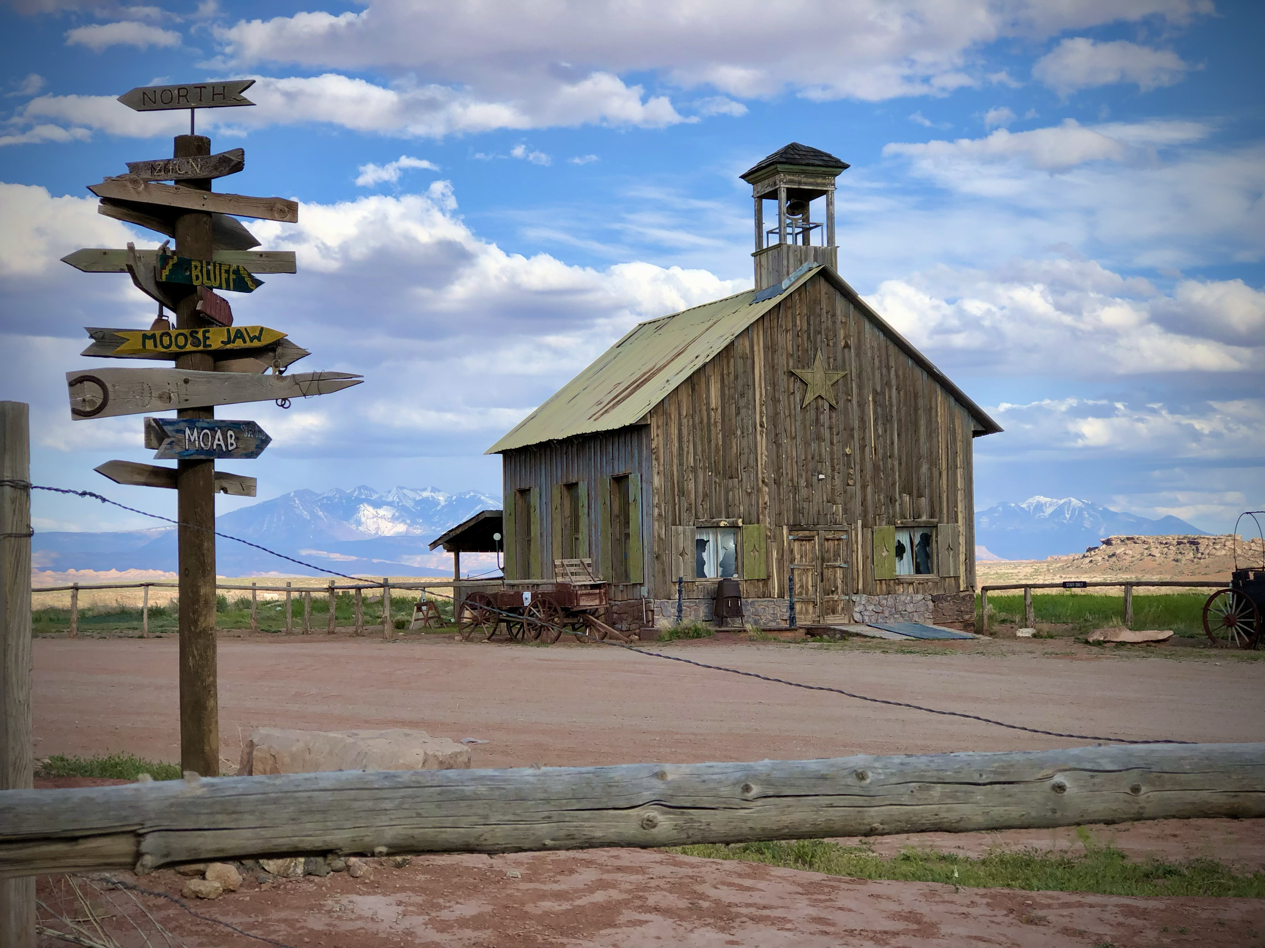 A rustic wooden building with a small bell tower, green shutters, and a star on the front, set against a backdrop of mountains and blue sky with clouds. A wooden fence surrounds the building, and a signpost with multiple arrows pointing in different 