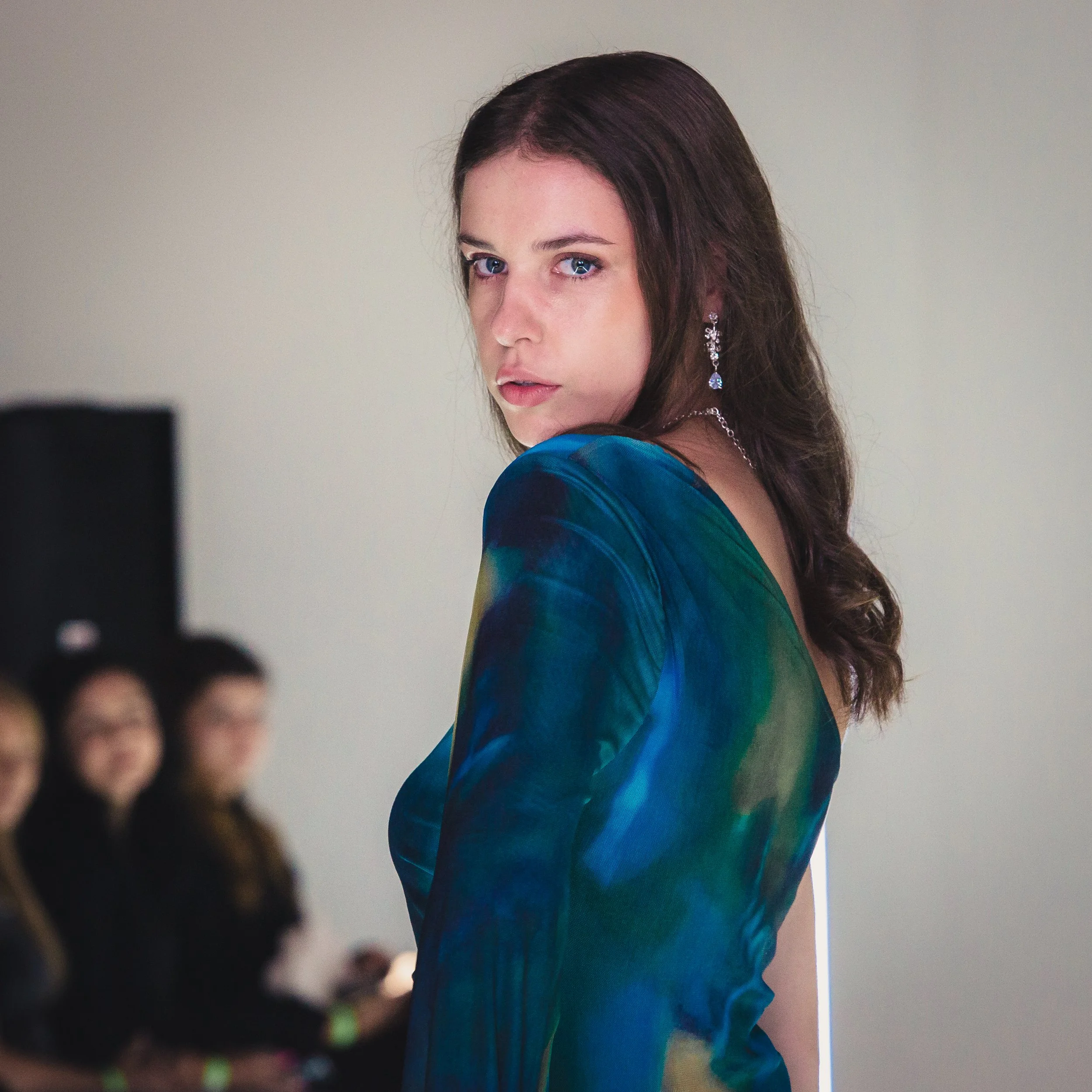 A woman with brown hair and blue eyes wearing a multicolored dress and dangling earrings. She is looking over her shoulder with a serious expression, blurred audience in the background.