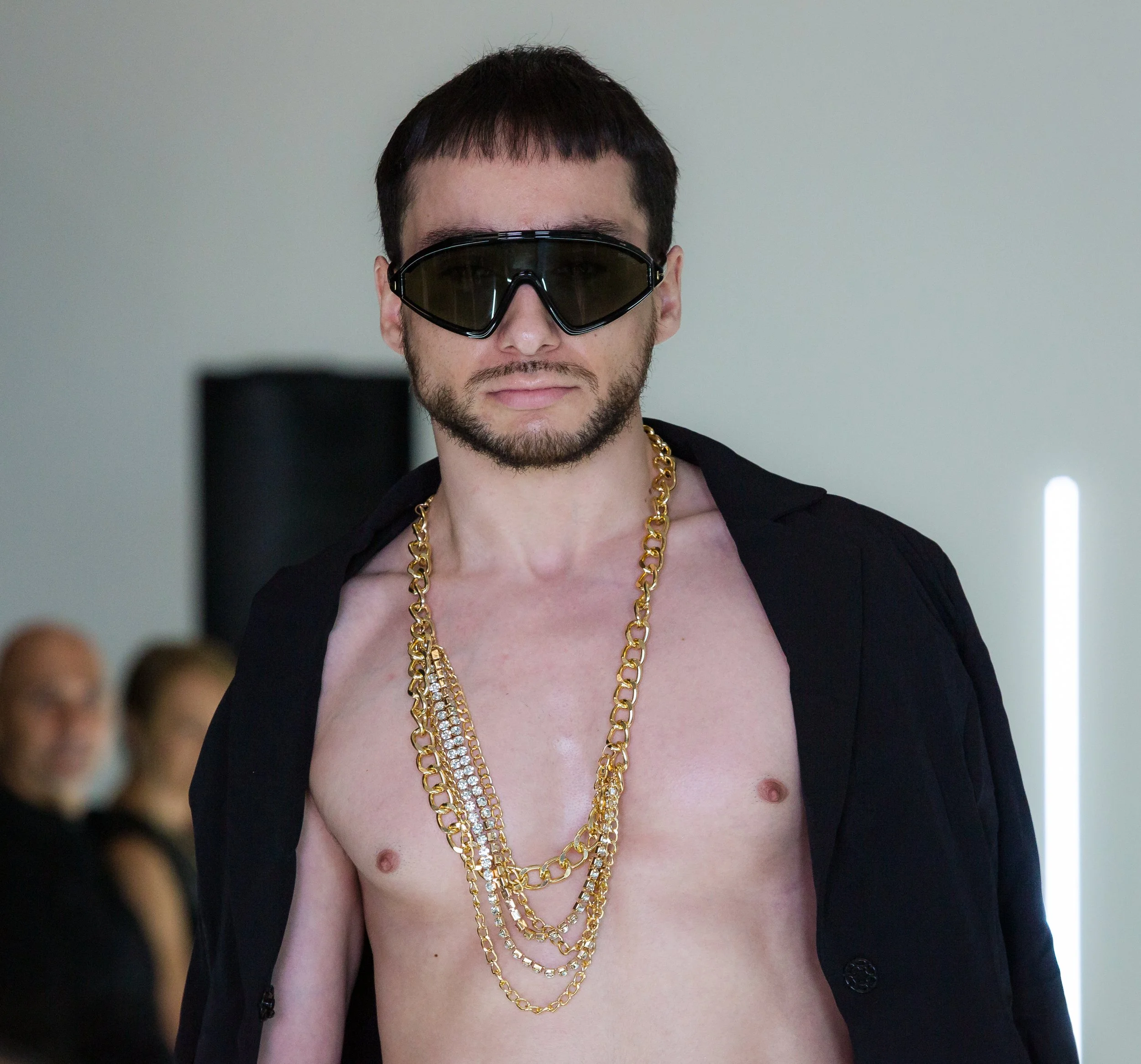 Man wearing sunglasses, a black jacket over his shoulders, and multiple gold chains around his neck, standing shirtless indoors.