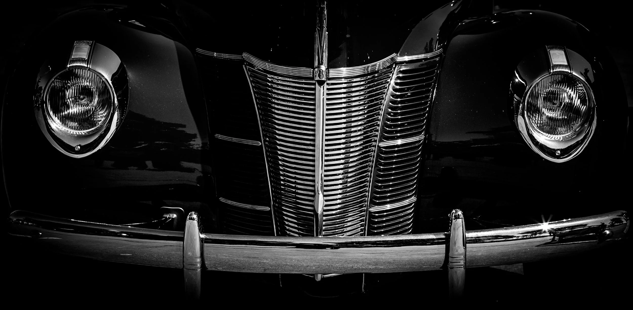 Front view of a vintage black car with a prominent chrome grille and round headlights, in black and white.