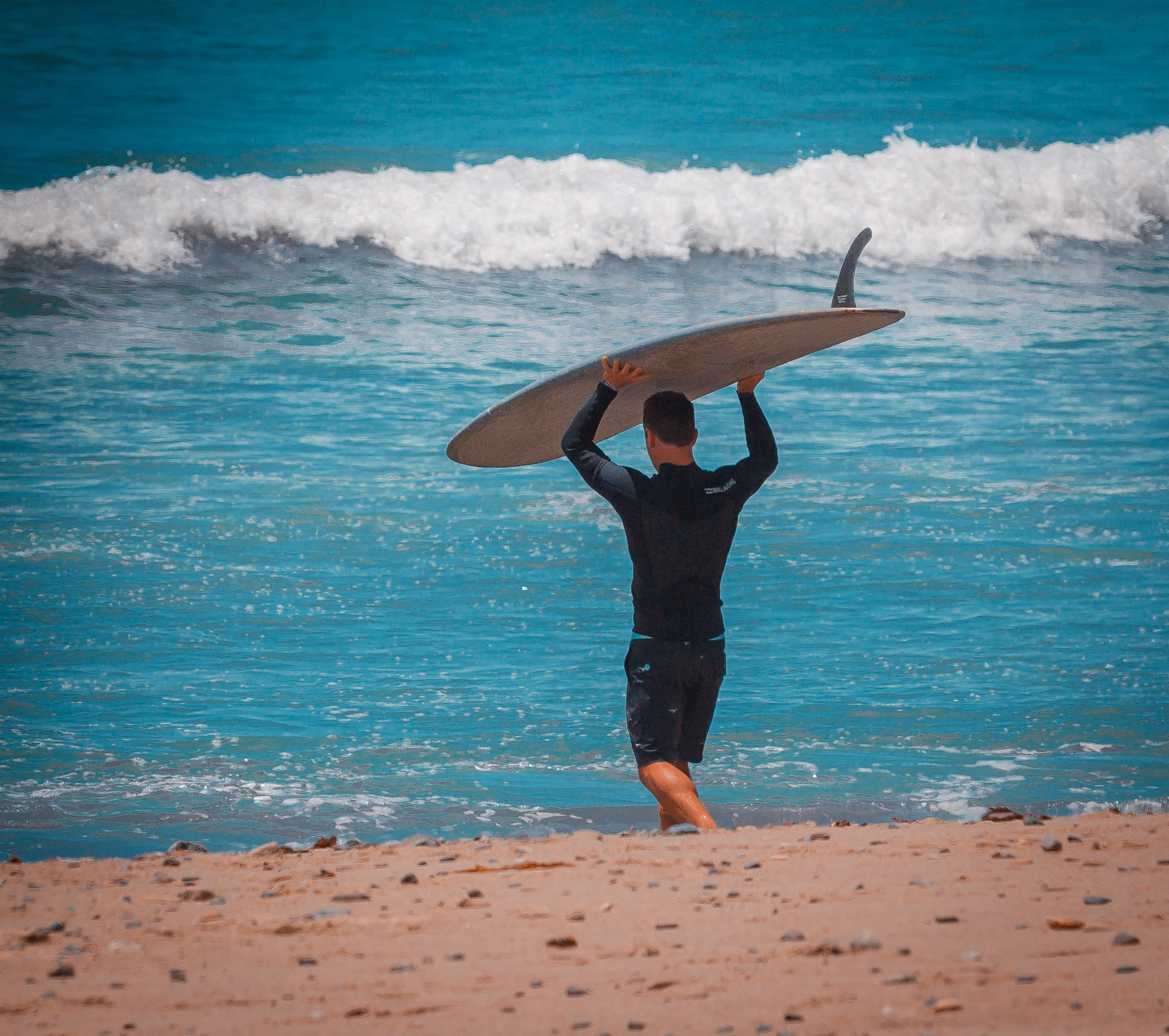 A man carrying a surfboard on his shoulders walking into the ocean at the beach.