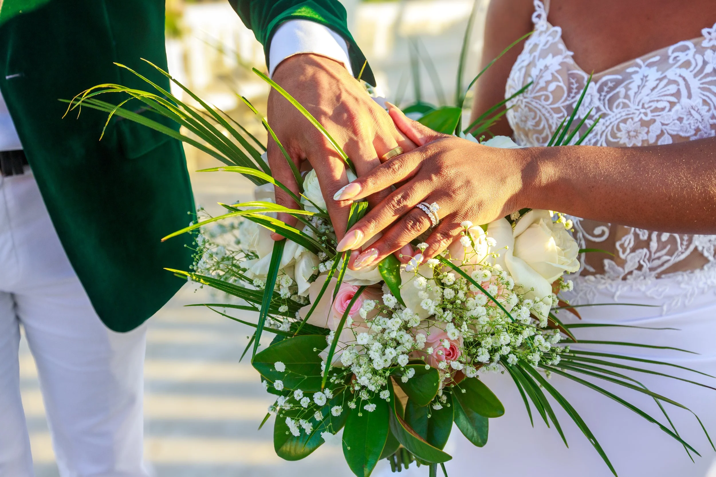 A close-up of a bride and groom holding hands over a bouquet of white and pink flowers and greenery during a wedding ceremony.
