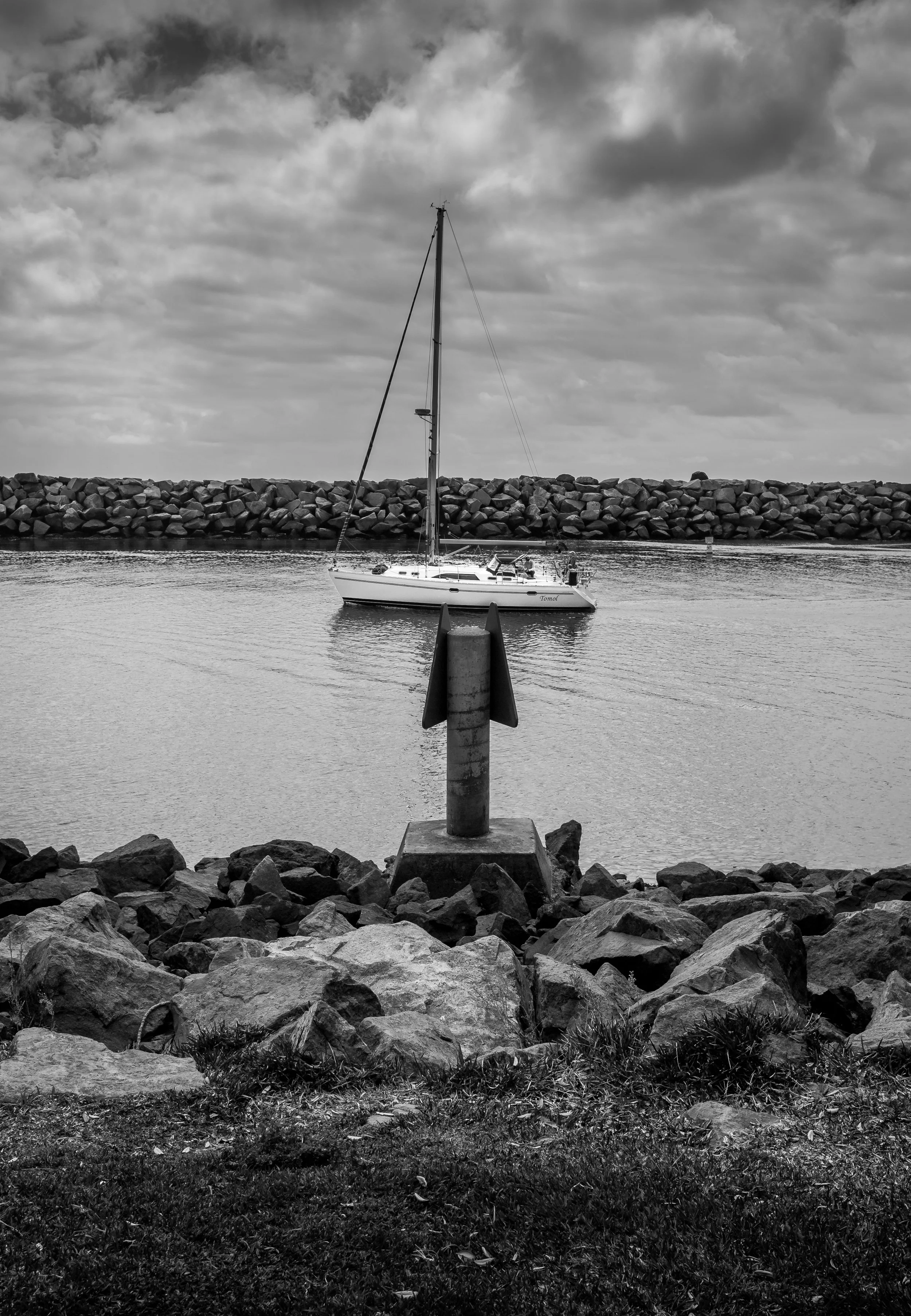 A black and white photo of a sailboat anchored in a marina, with a rocky breakwater in the background and a large arrow-shaped navigation marker in the foreground.