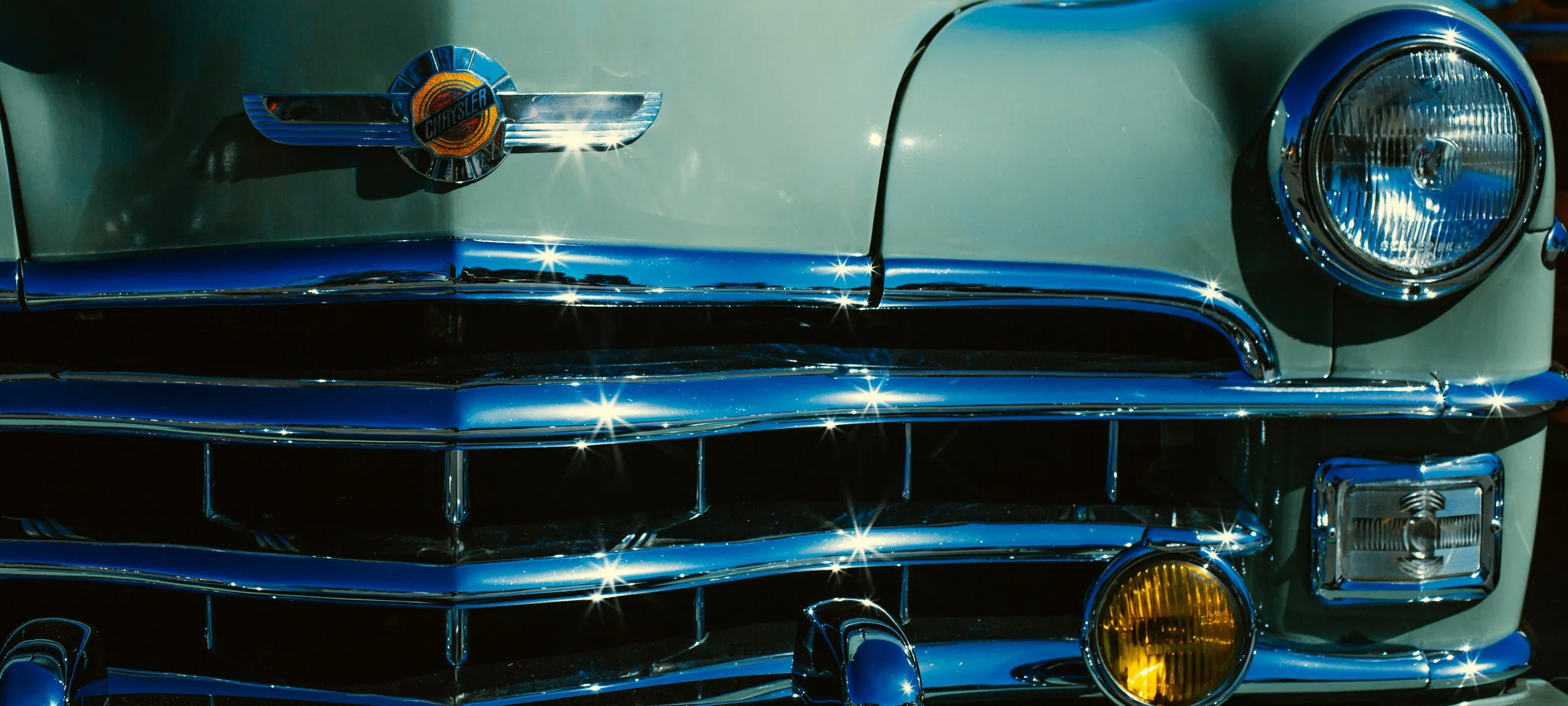 Close-up of the front of a vintage Chrysler car, featuring chrome details, a round headlight, and a Chrysler badge on the hood.