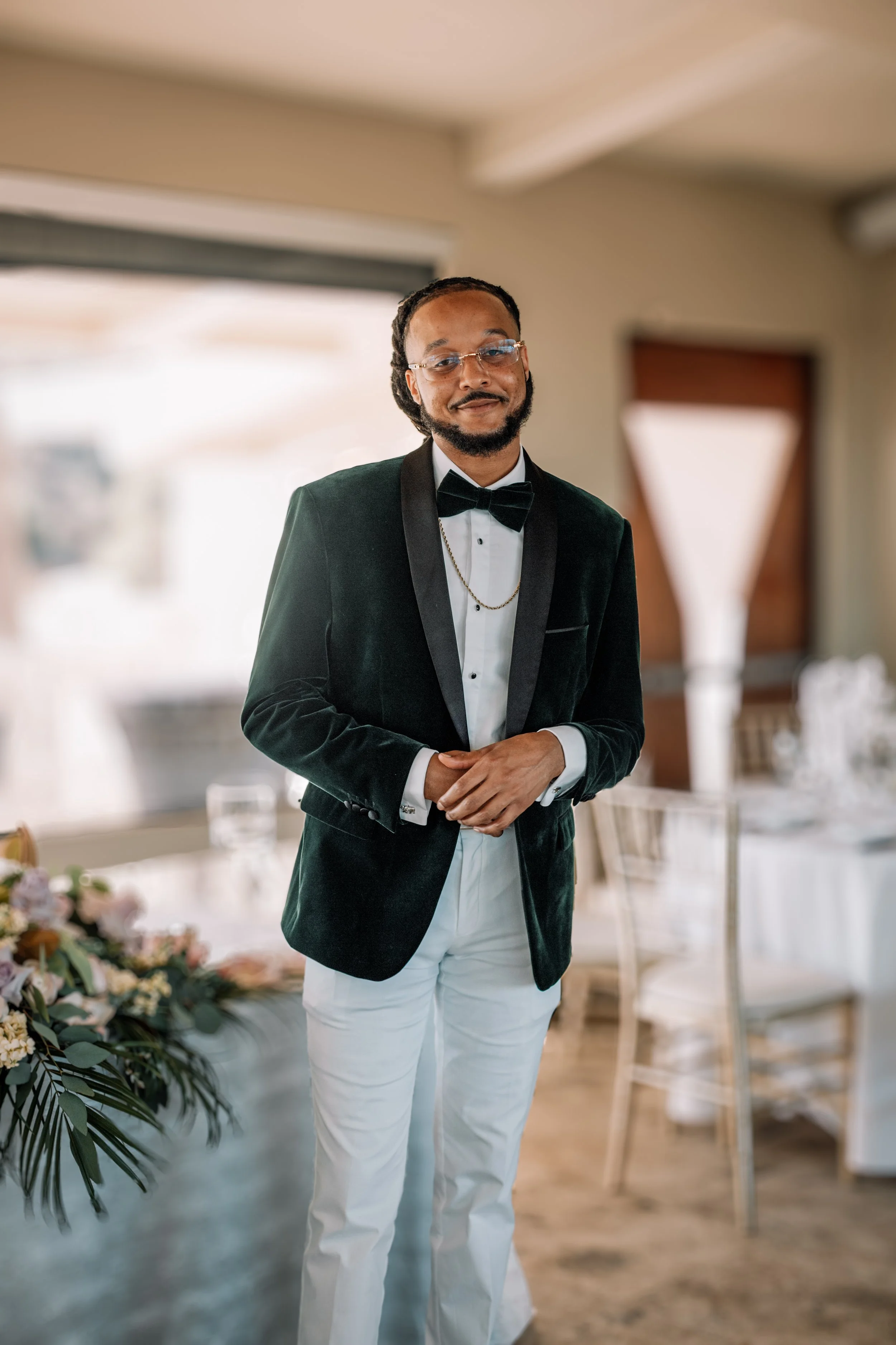 A man dressed in a tuxedo with a black velvet blazer, white shirt, black bow tie, and white pants, standing in a well-lit indoor space with dining tables and floral arrangements.