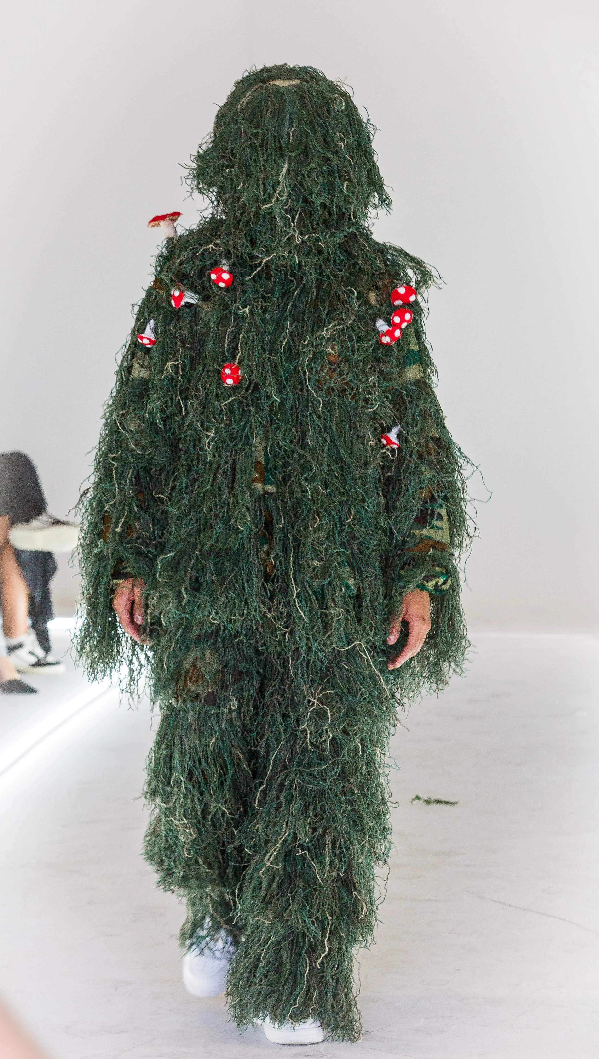 Person dressed in a green, textured, camouflage-like outfit resembling a ghillie suit with red and white mushroom decorations.