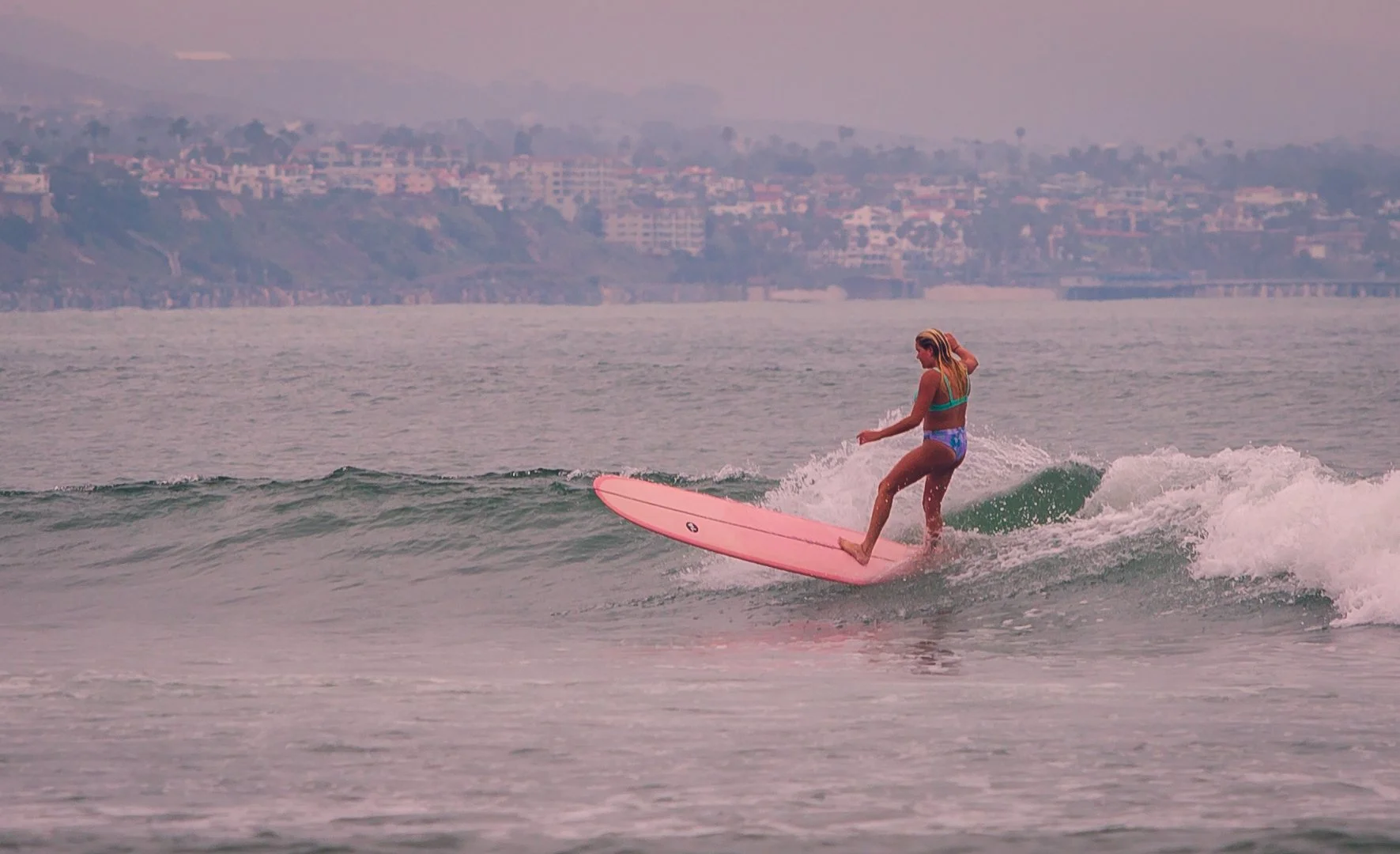 A woman surfing on a pink surfboard in the ocean near a city coastline during dusk or dawn.