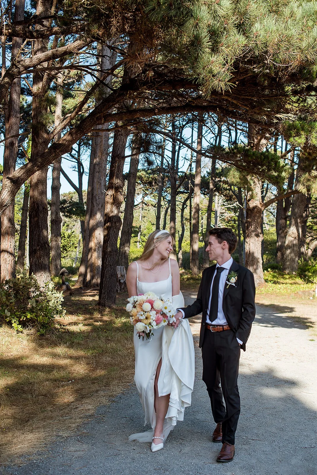 A bride and groom holding hands and smiling at each other in a forest setting. The bride wears a white wedding dress and carries a bouquet of flowers. The groom wears a black suit with a white shirt and tie.