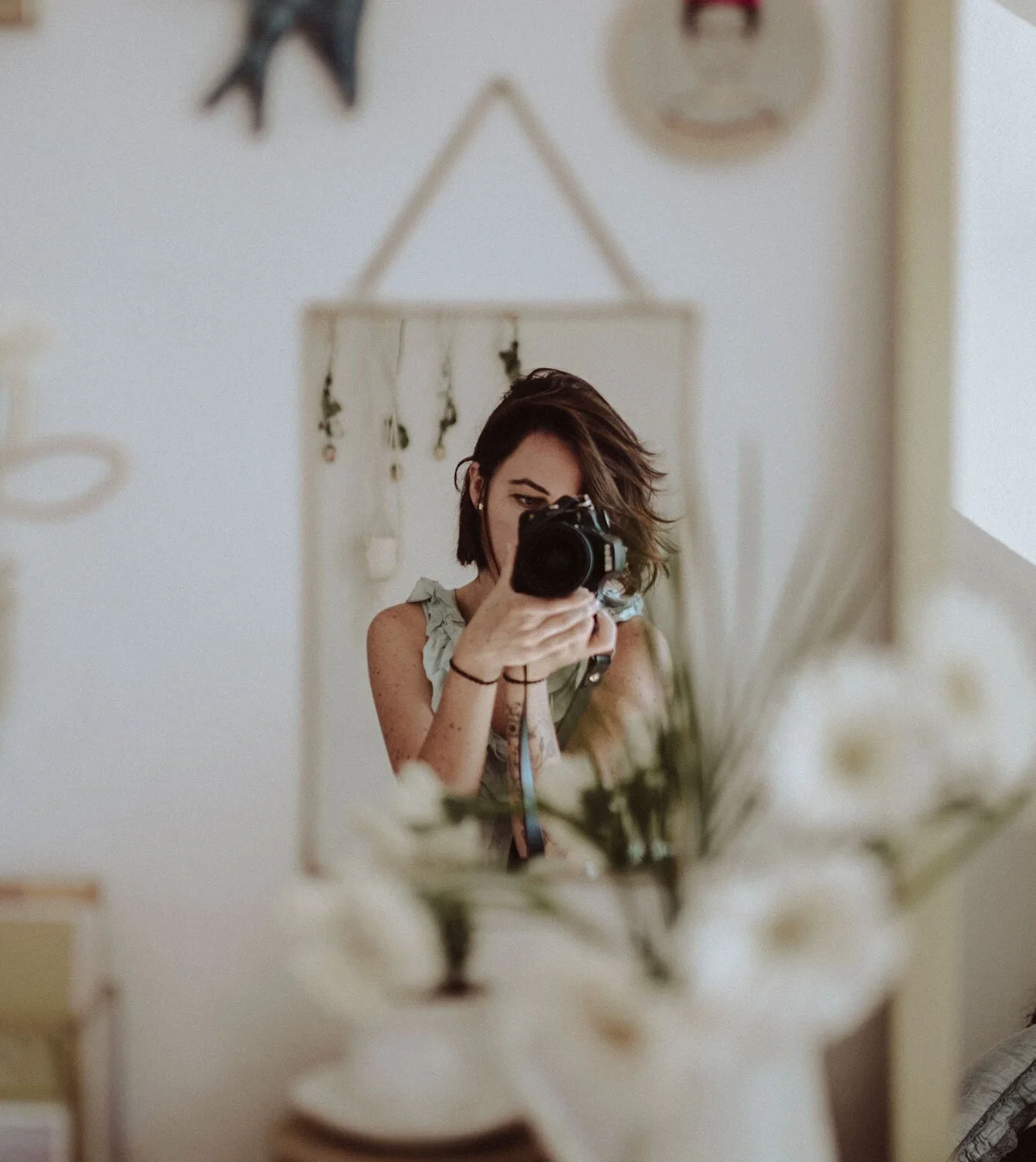Woman taking a mirror selfie indoors, holding a camera, with flowers in foreground and decorative wall hangings behind.