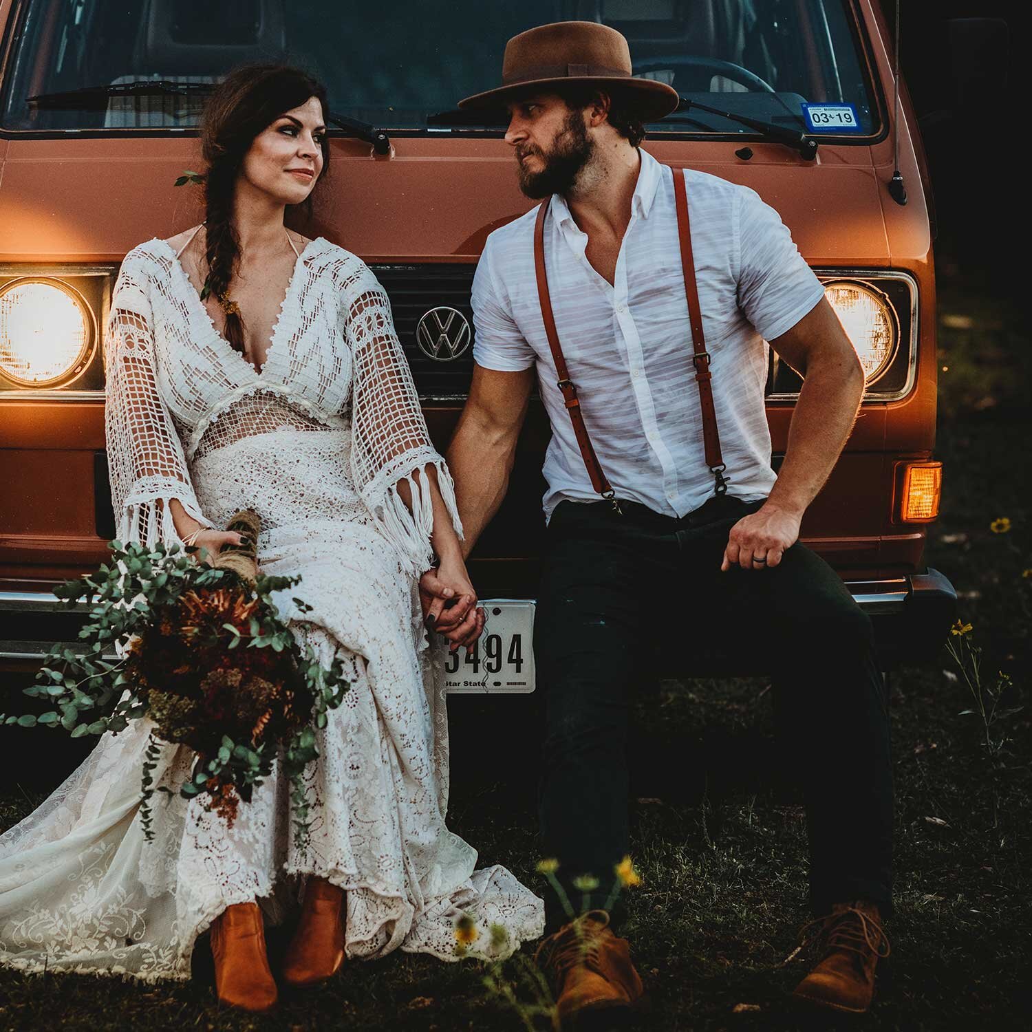 A couple sitting in front of an orange Volkswagen van during sunset, holding hands. The woman is wearing a bohemian lace wedding dress, ankle boots, and is holding a bouquet of flowers. The man is dressed casually with a white shirt, suspenders, and a wide-brimmed hat.