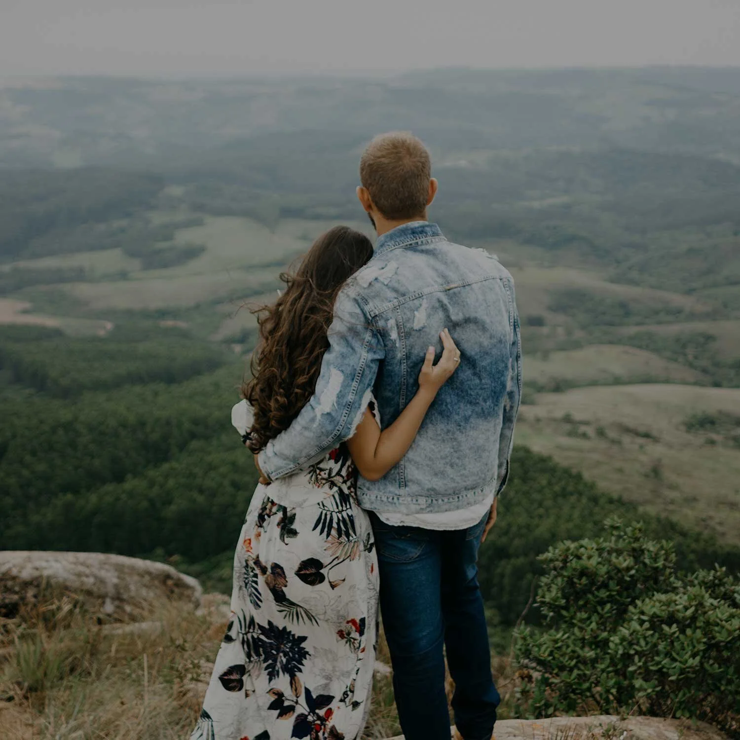 A couple standing on a hilltop overlooking a scenic valley, with the woman embracing the man from the side.