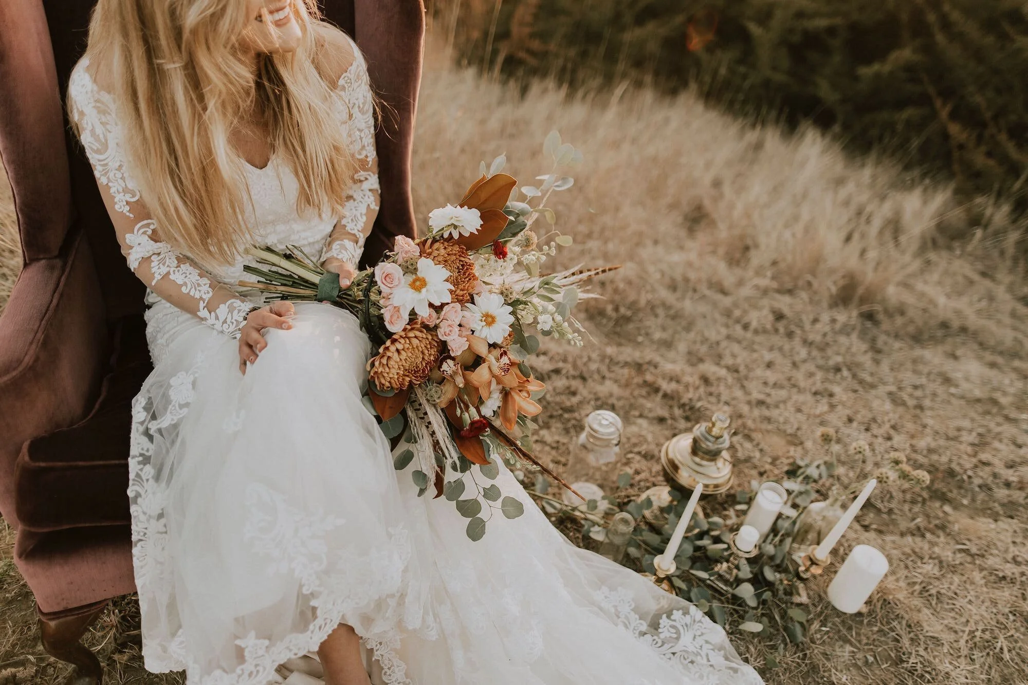 Bride sitting on a vintage armchair outdoors, wearing a lace wedding dress, holding a bouquet of flowers, next to candles and greenery.