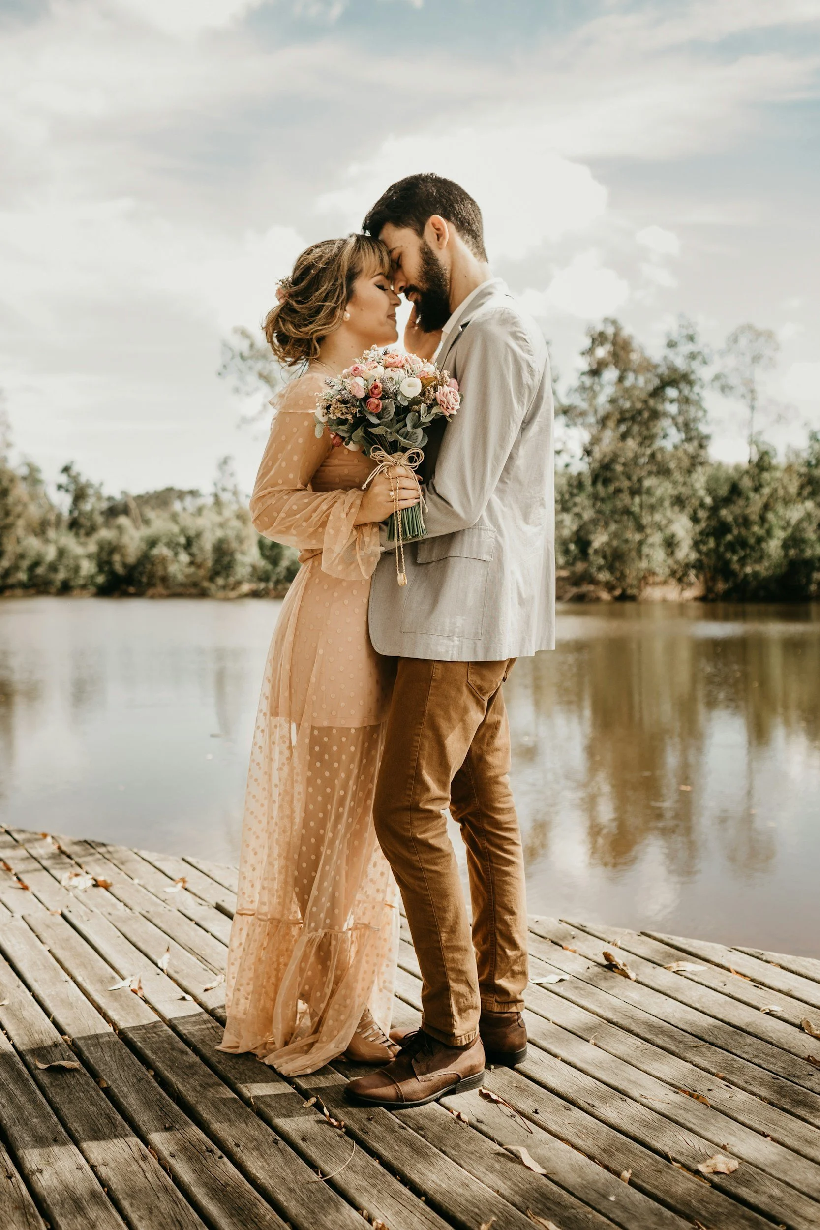 A couple embraces on a wooden dock by a river, with trees in the background, during daylight. The woman is holding a bouquet of flowers and wearing a beige, dotted dress, while the man wears a light gray blazer and brown pants.