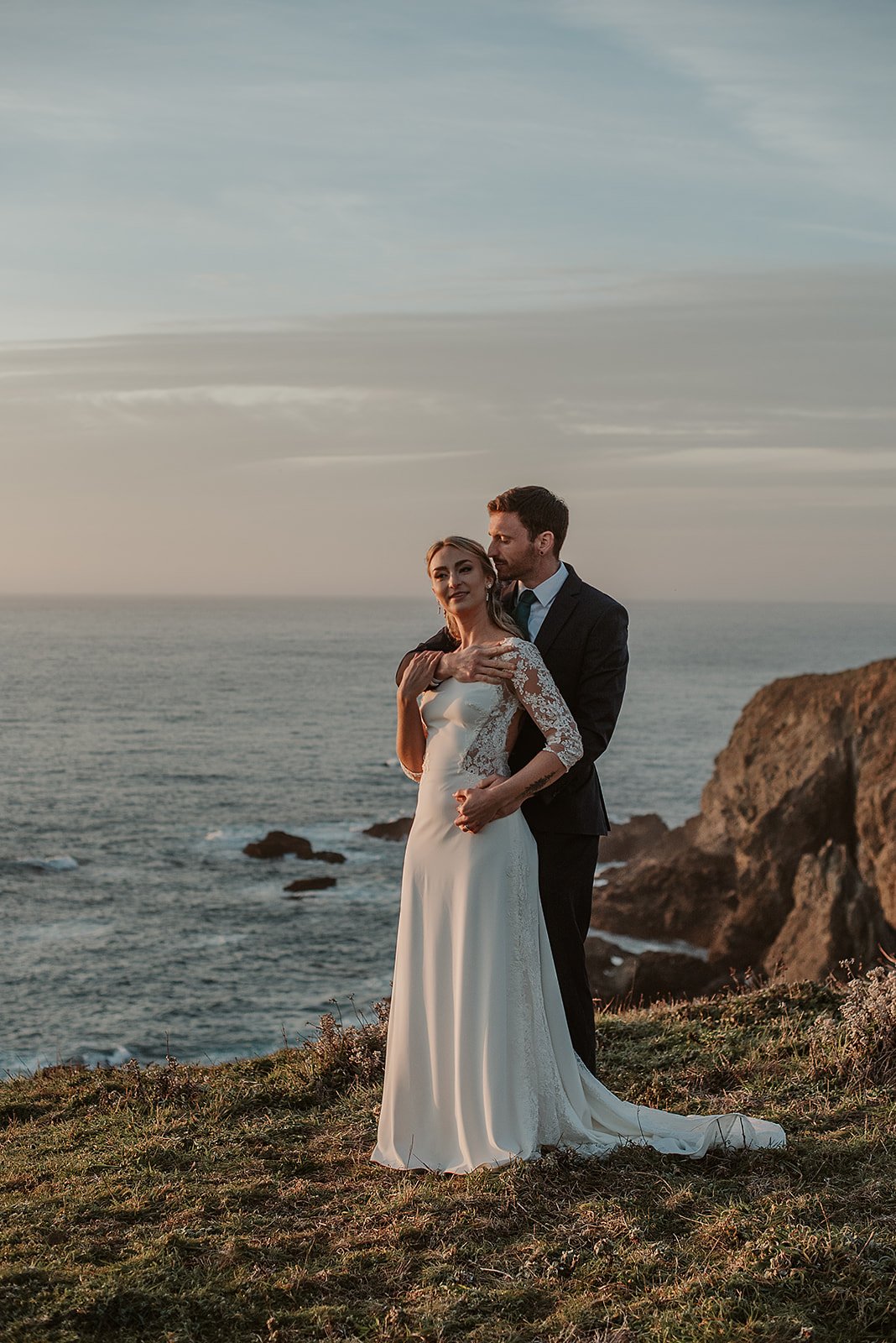 A bride and groom standing on a grassy cliff with the ocean in the background, during sunset.