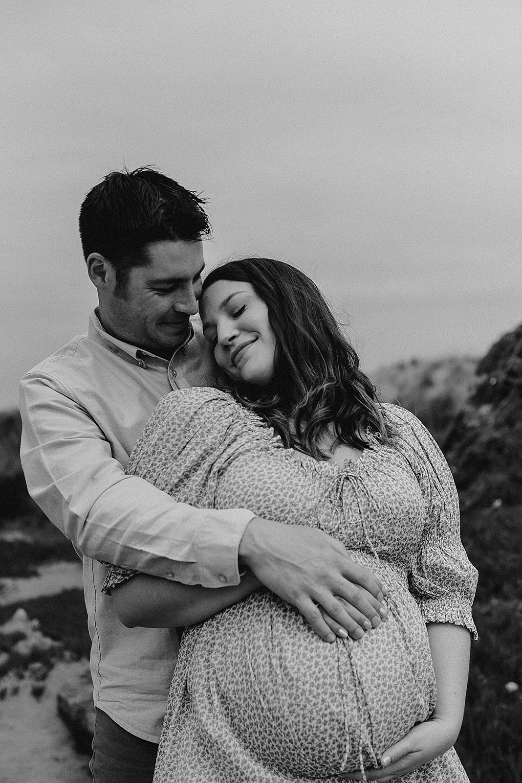 A loving couple embraces outdoors, with the woman visibly pregnant, both smiling gently with eyes closed.