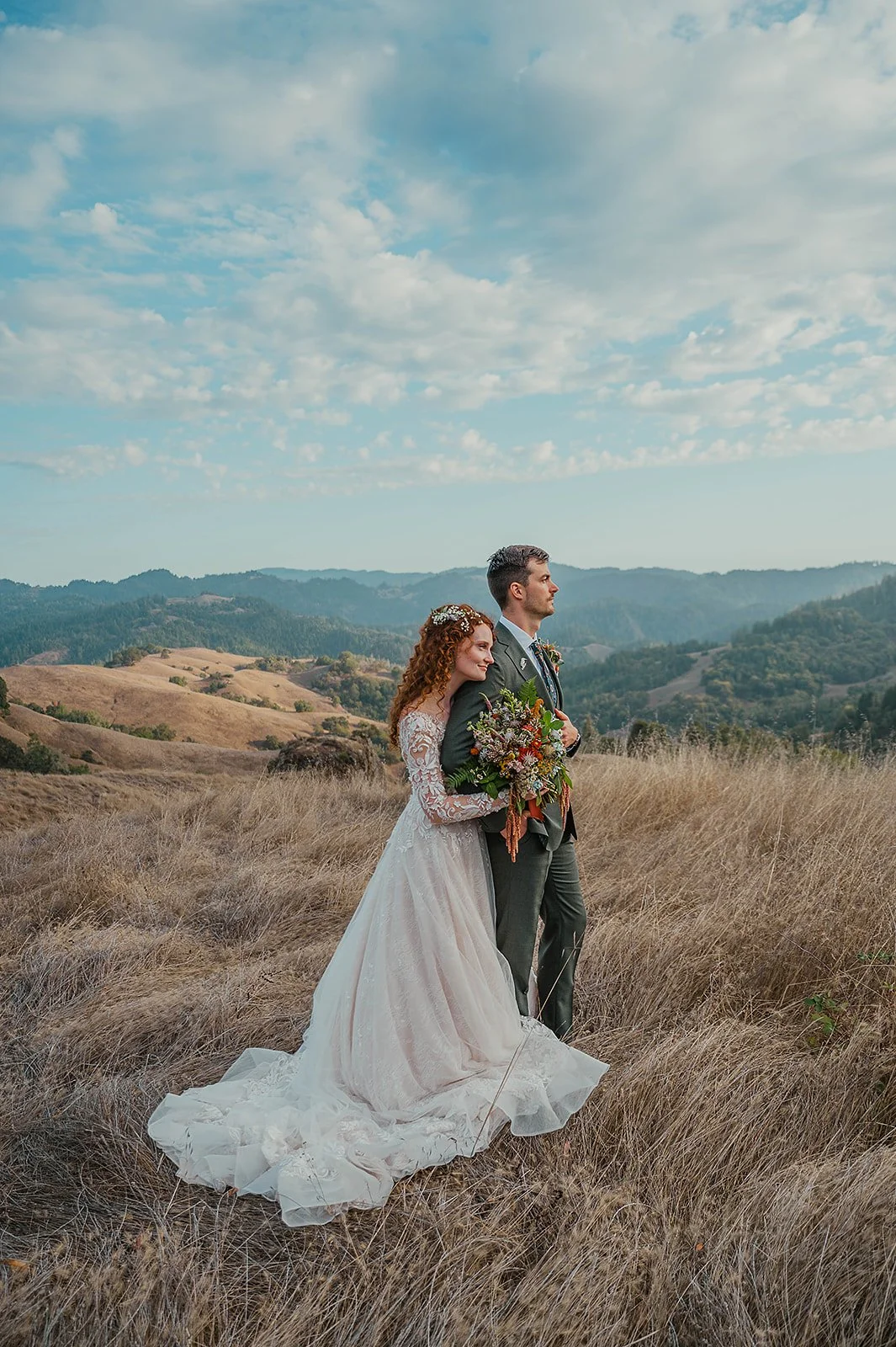 A bride and groom standing in a grassy field with rolling hills and a partly cloudy sky in the background. The bride is holding a bouquet of flowers and leaning her head against the groom's shoulder.