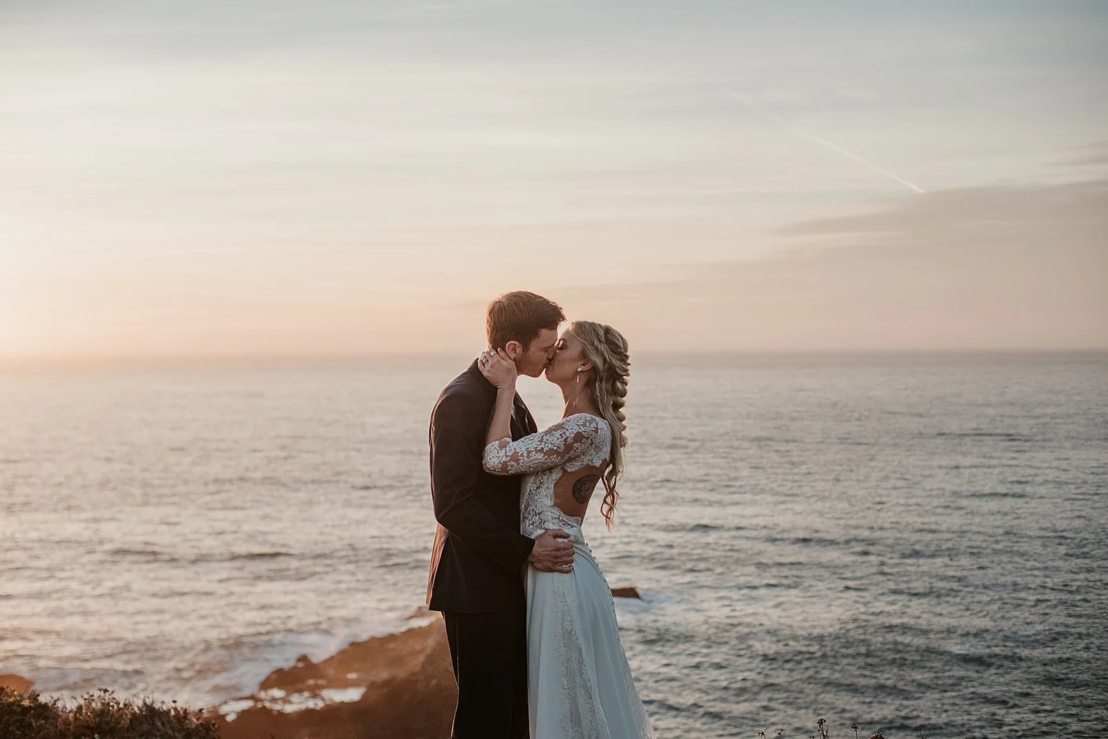 A couple in wedding attire sharing a kiss by the ocean at sunset.