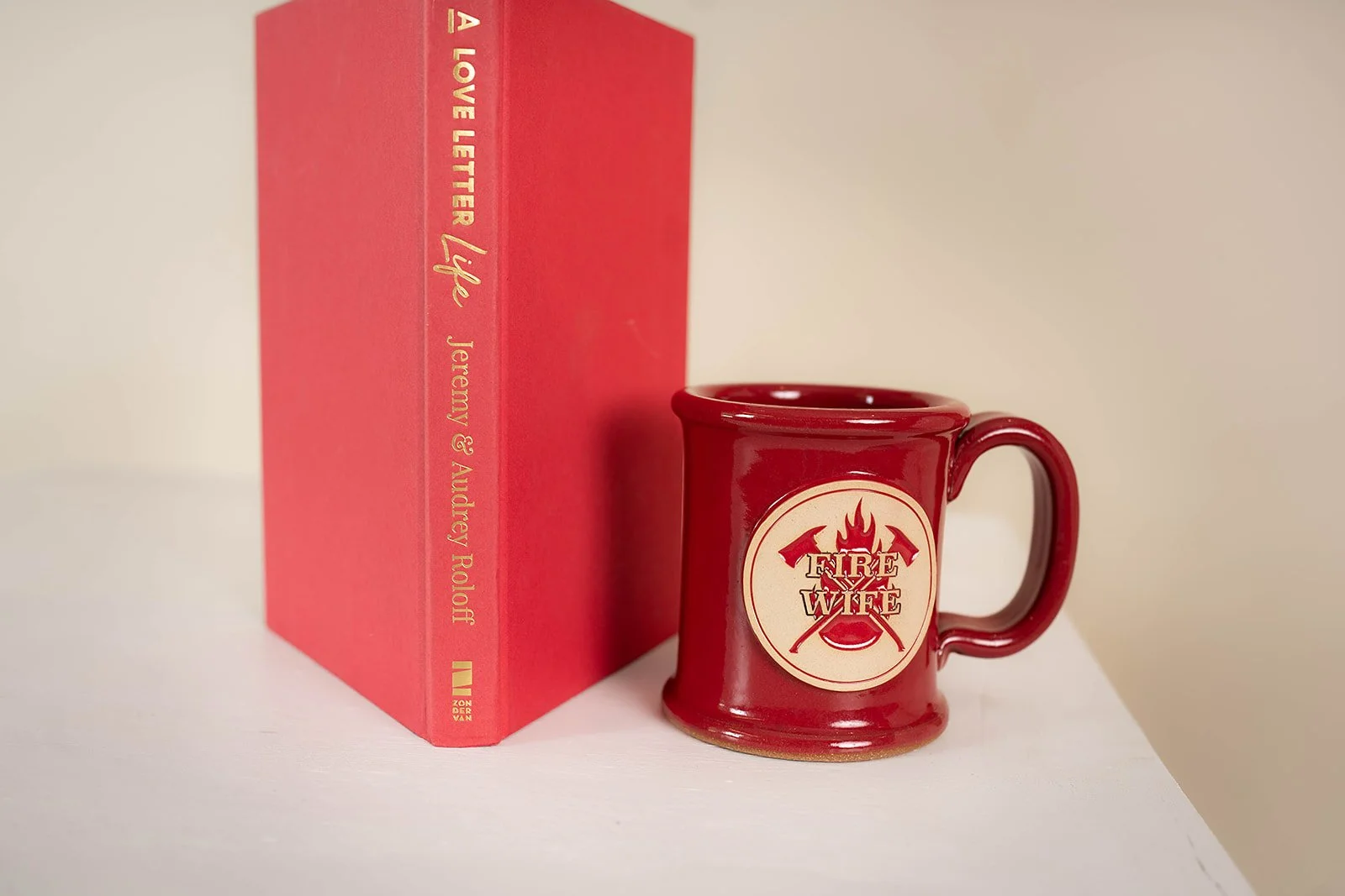 A red hardcover book titled "A Love Letter Life" by Jeramy & Audrey Poole standing next to a red ceramic mug with the words "FIRE WIFE" and images of crossed axes and flames on it, placed on a white surface against a beige background.