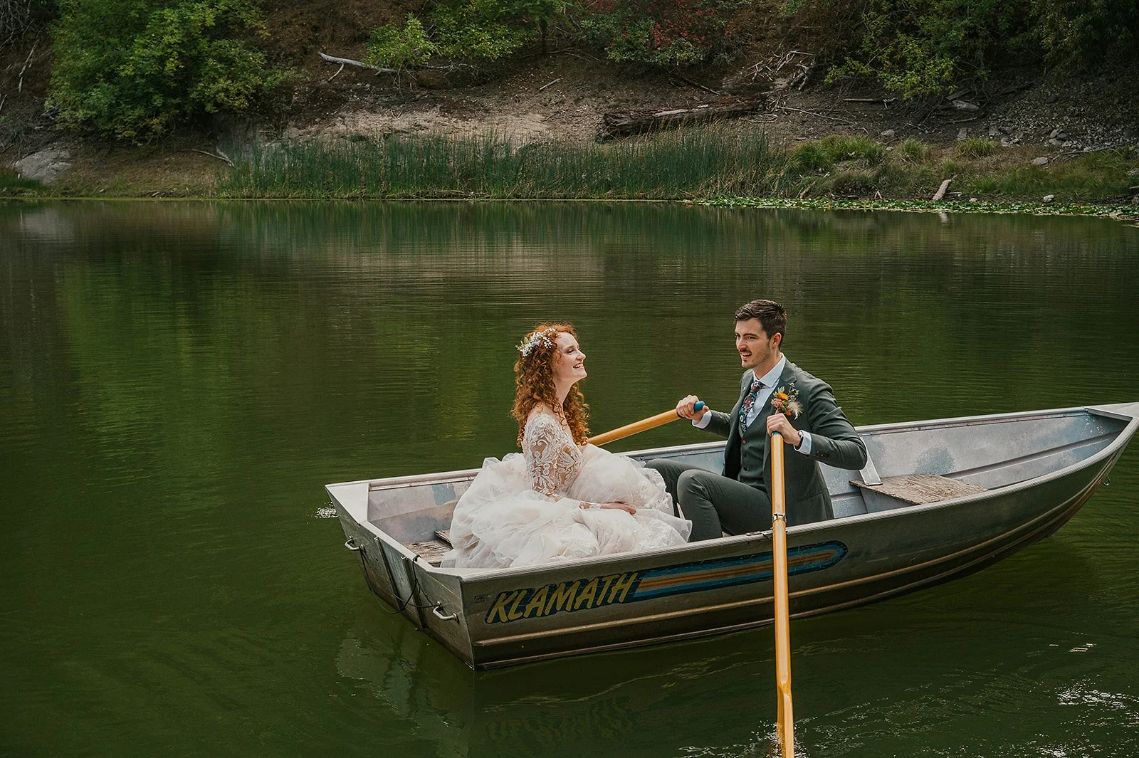 A bride and groom in wedding attire enjoying a rowboat on a calm lake, surrounded by greenery.