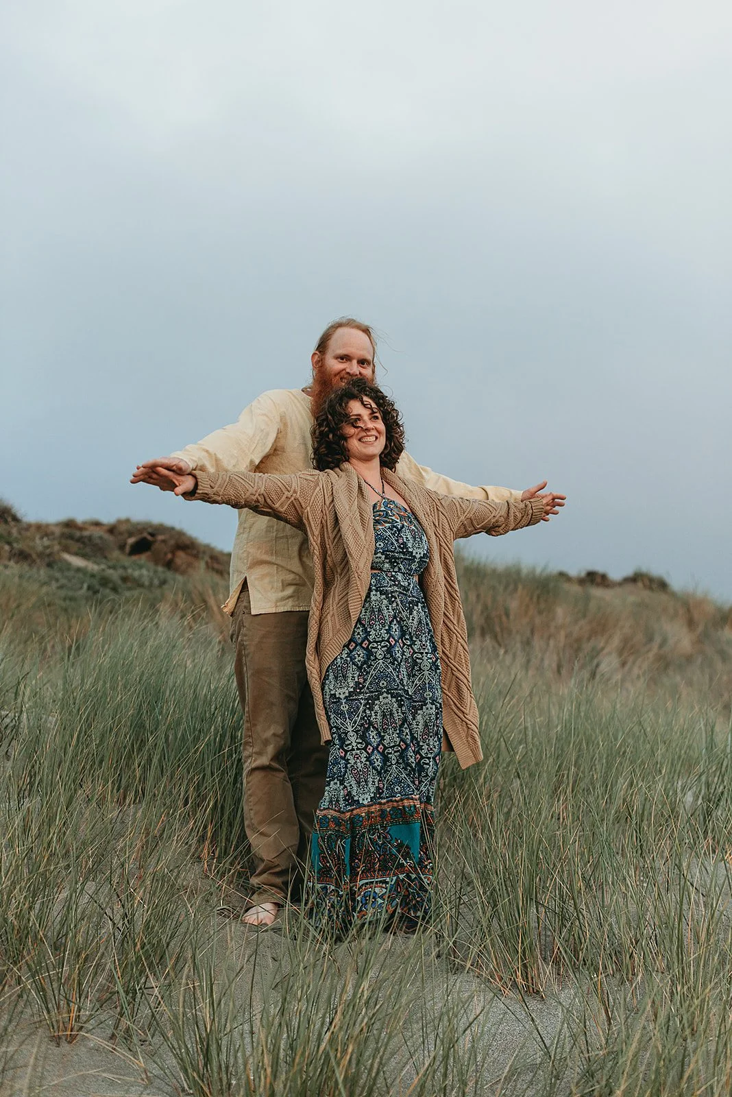 A couple standing in a grassy field, smiling, with arms outstretched, under a cloudy sky.