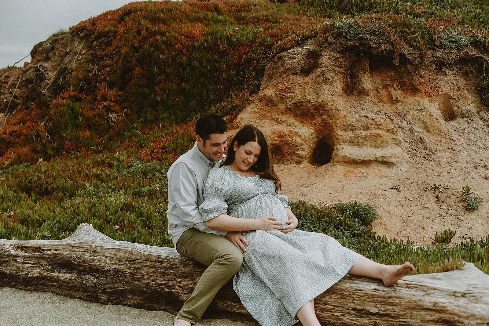 A couple sitting on a log near the beach, with the woman holding her pregnant belly and the man embracing her from behind, surrounded by sand and coastal vegetation.