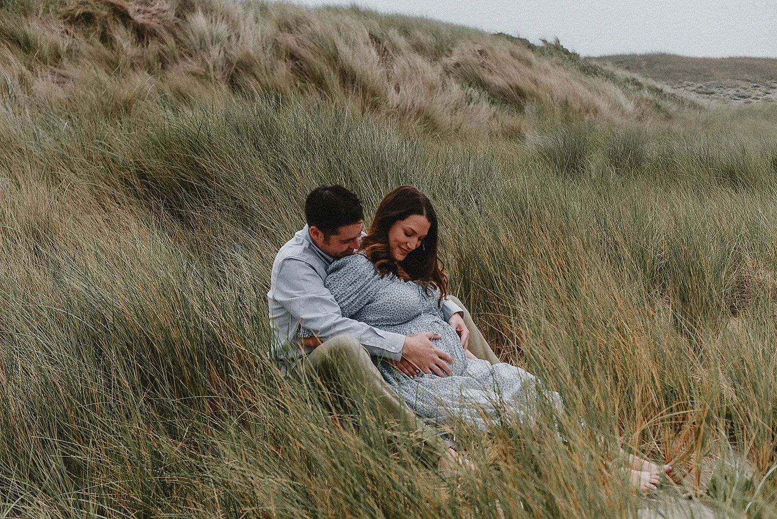 A couple sitting in tall grass on a hillside, with the man embracing the pregnant woman from behind, both smiling and looking down at her belly.