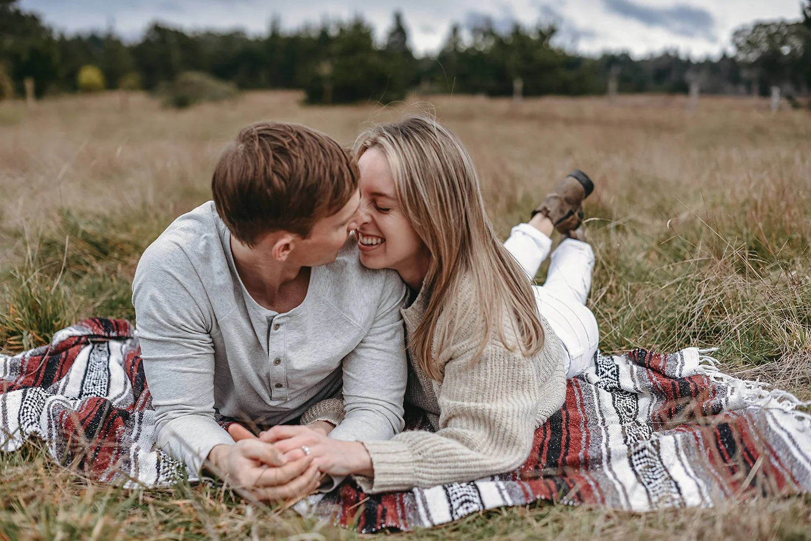 A couple lying on a blanket in a field, smiling as they touch noses.