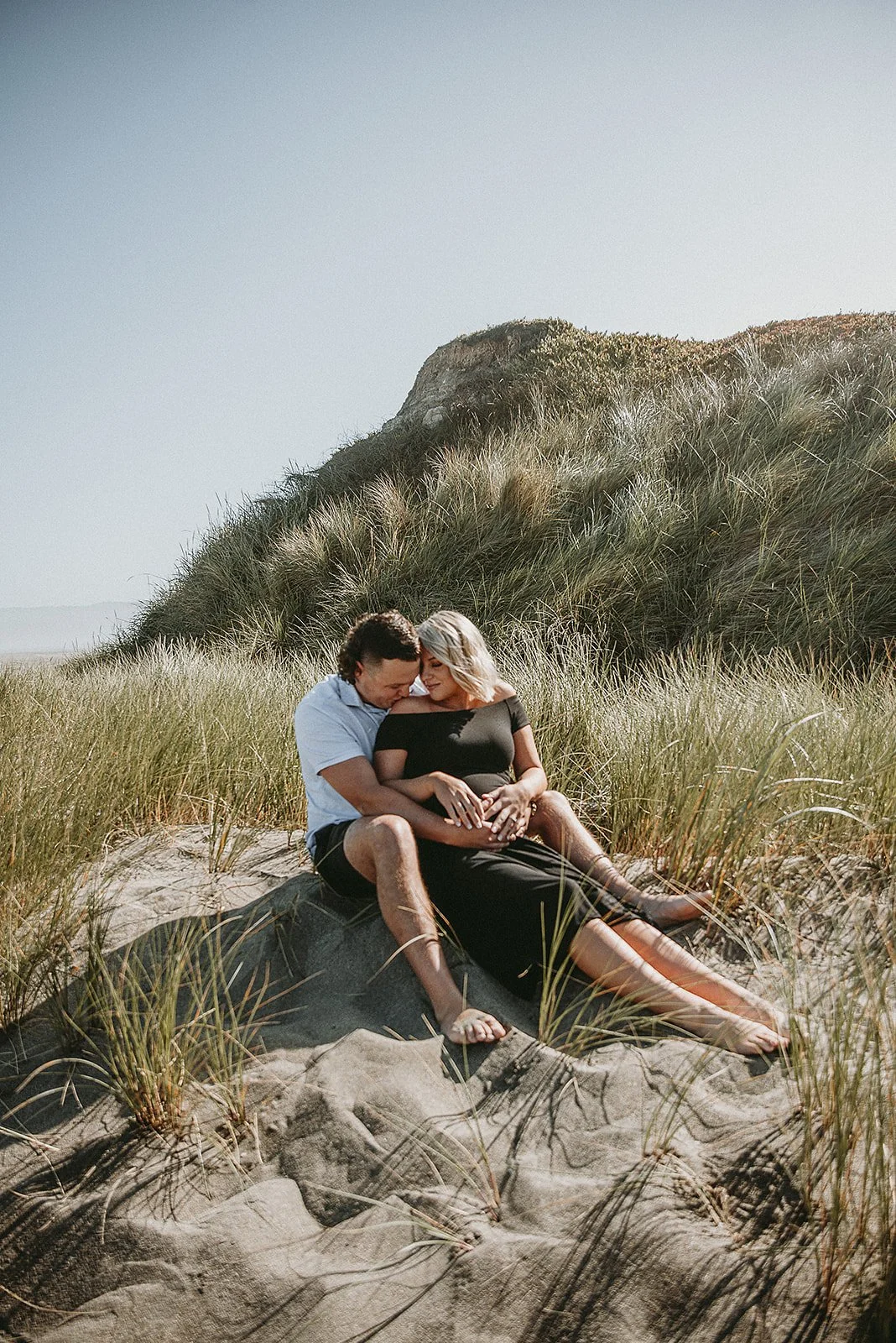 A couple sitting closely together on a sandy beach surrounded by grass and dunes, sharing an intimate moment in natural sunlight.