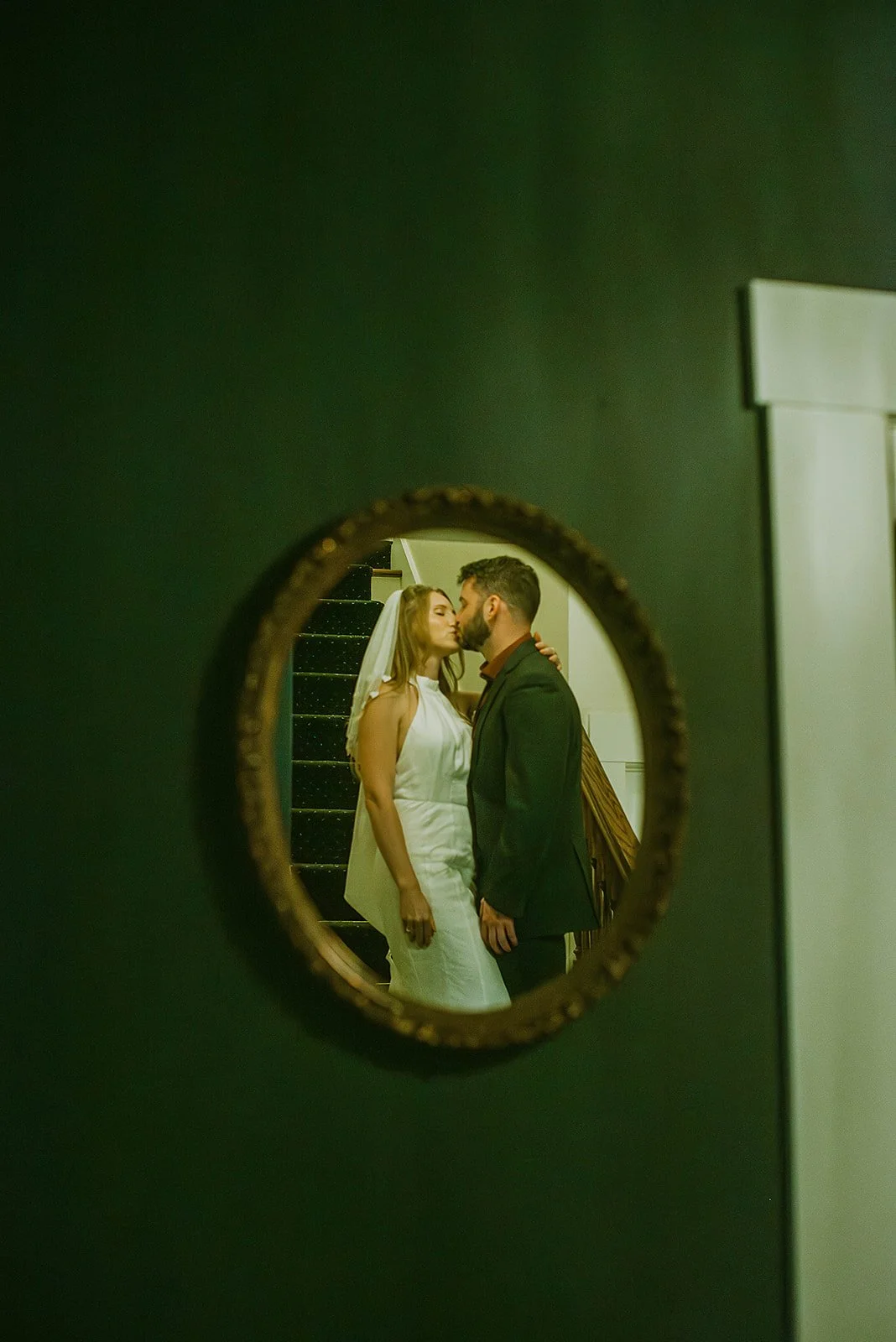 A bride and groom kissing, seen through a small round mirror mounted on a green wall, with a staircase visible in the background.