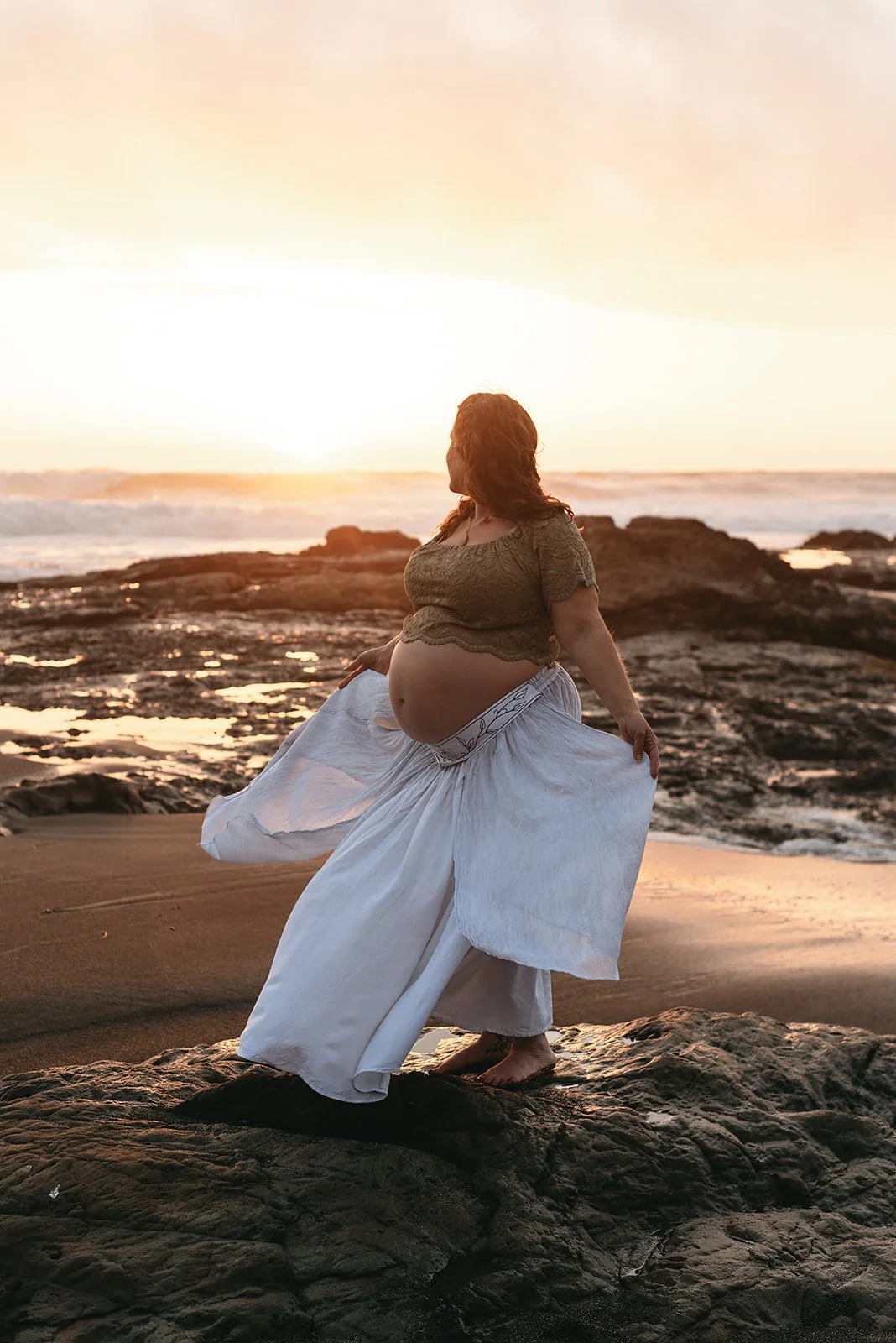 Pregnant woman standing on rocks at the beach during sunset, wearing a gold top and white wide-leg pants, holding the edges of her pants.