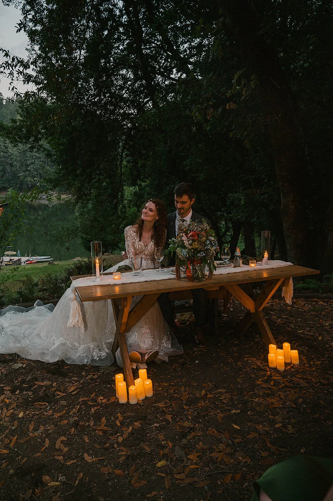 A bride and groom sit at a rustic outdoor dinner table surrounded by candles and a floral centerpiece, by a lakeside, beneath trees at dusk.