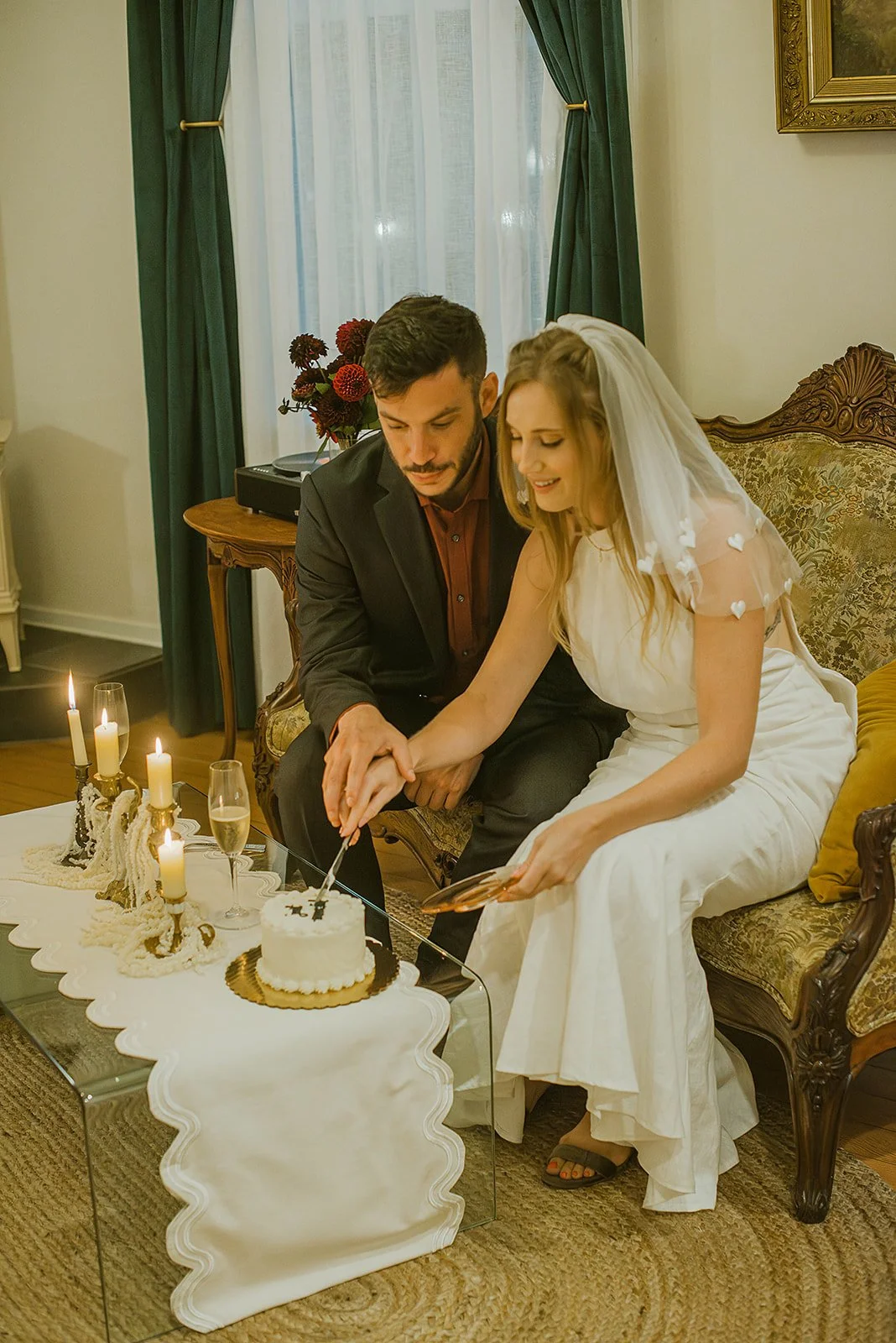 A newlywed couple sitting on a vintage sofa cutting a wedding cake together during their celebration, surrounded by candles and flowers in a warmly decorated room.