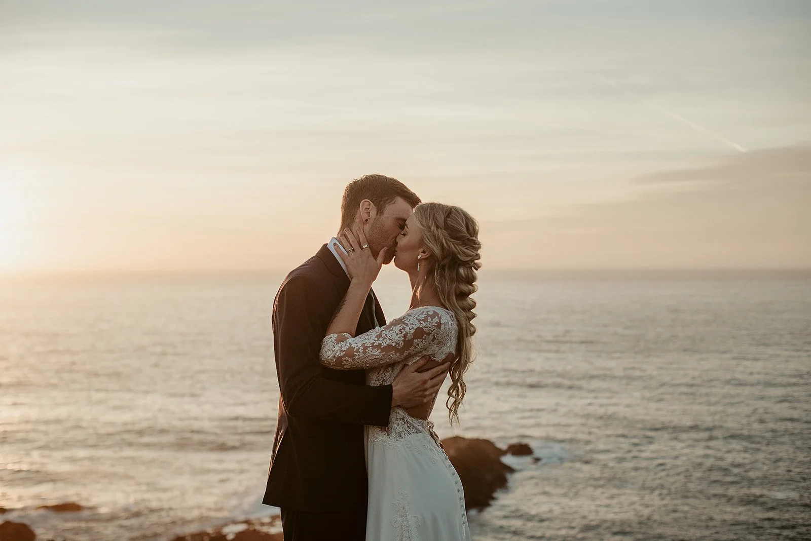 A couple in wedding attire sharing a kiss on a beach at sunset.