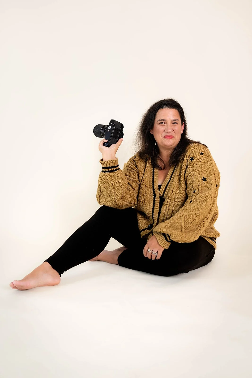 A woman sitting on the floor holding a camera in her right hand, wearing a mustard yellow sweater with black stars and black pants, making a humorous face against a plain light background.