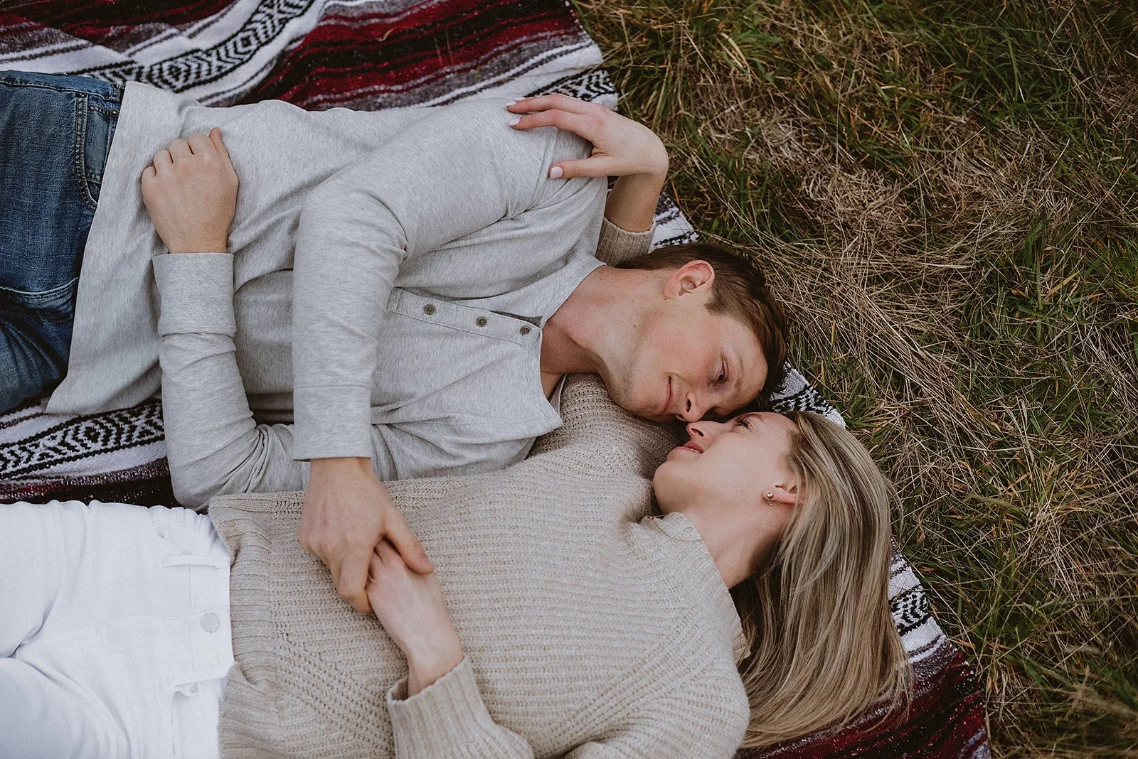 A young couple lying on the ground on a blanket in a grassy area, gazing at each other lovingly.